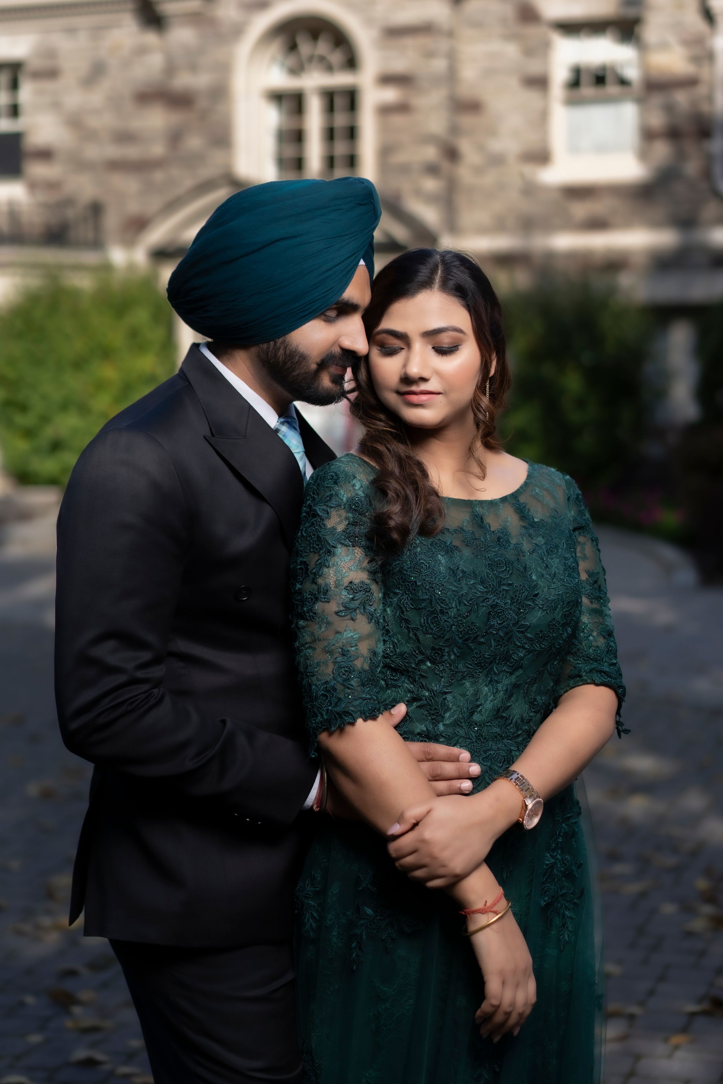 Elegant couple in formal attire poses against stone building with autumn leaves.