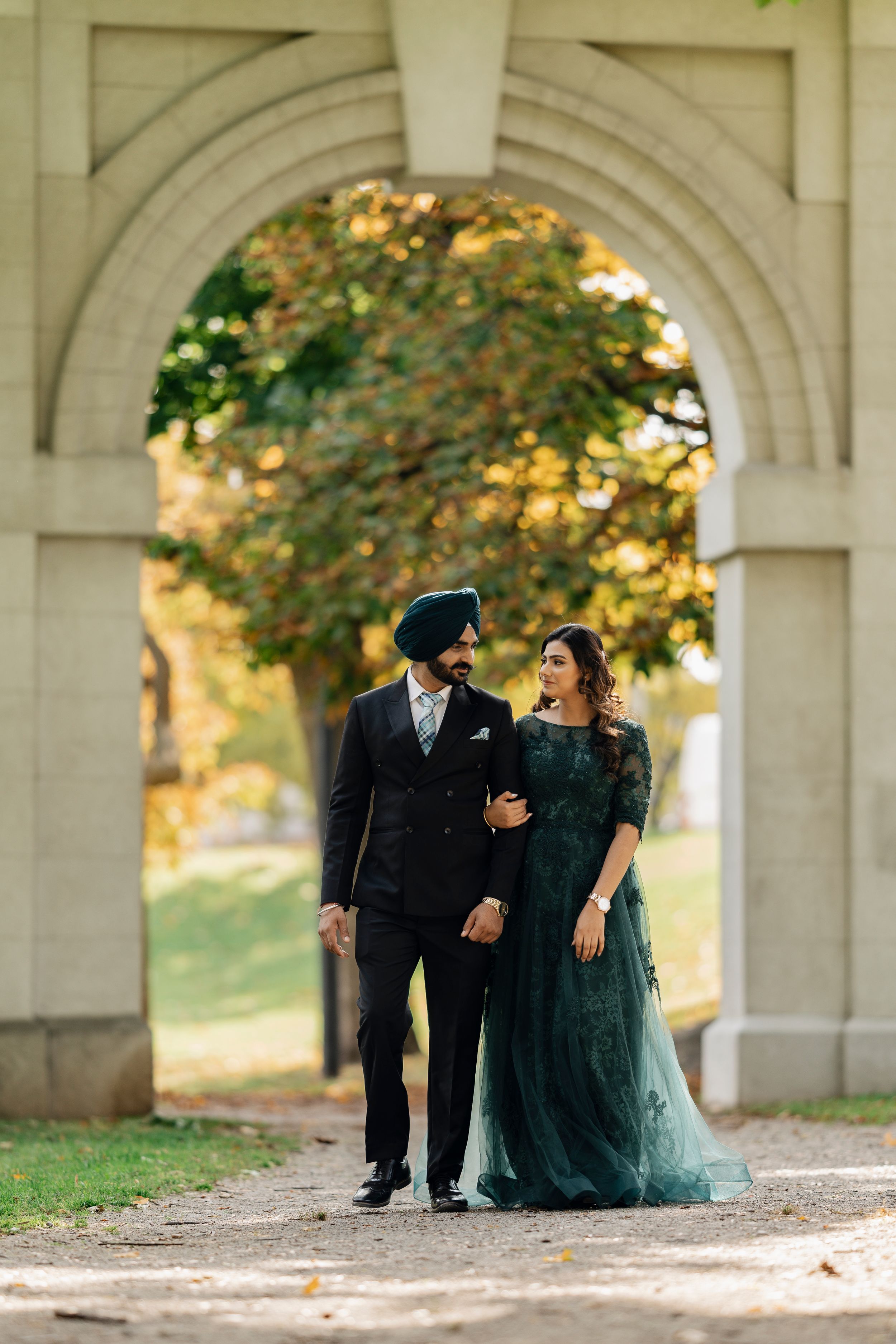 An elegant couple walks beneath a grand archway during autumn.