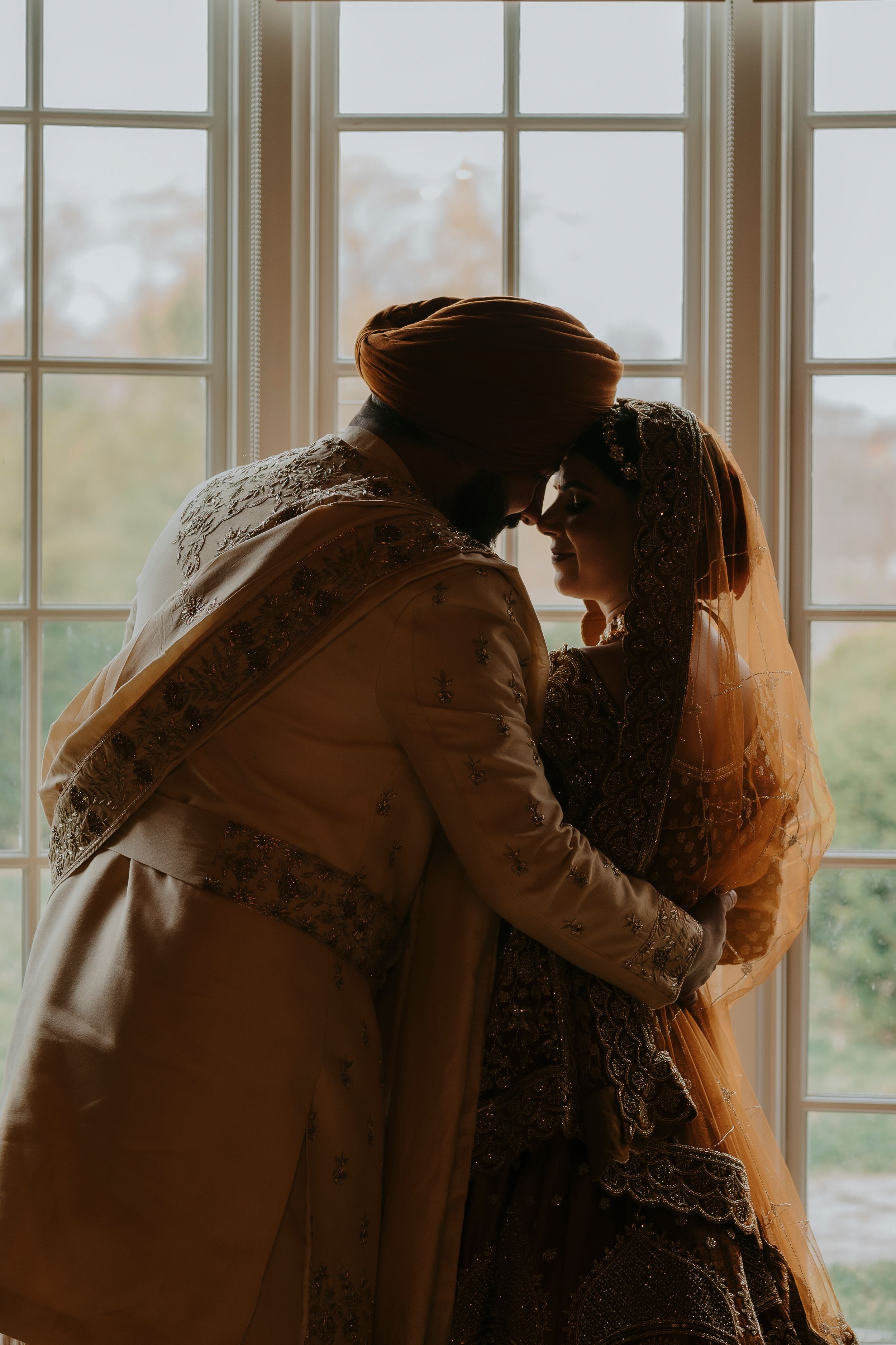 A silhouetted couple in traditional attire shares an intimate moment by a sunlit window.