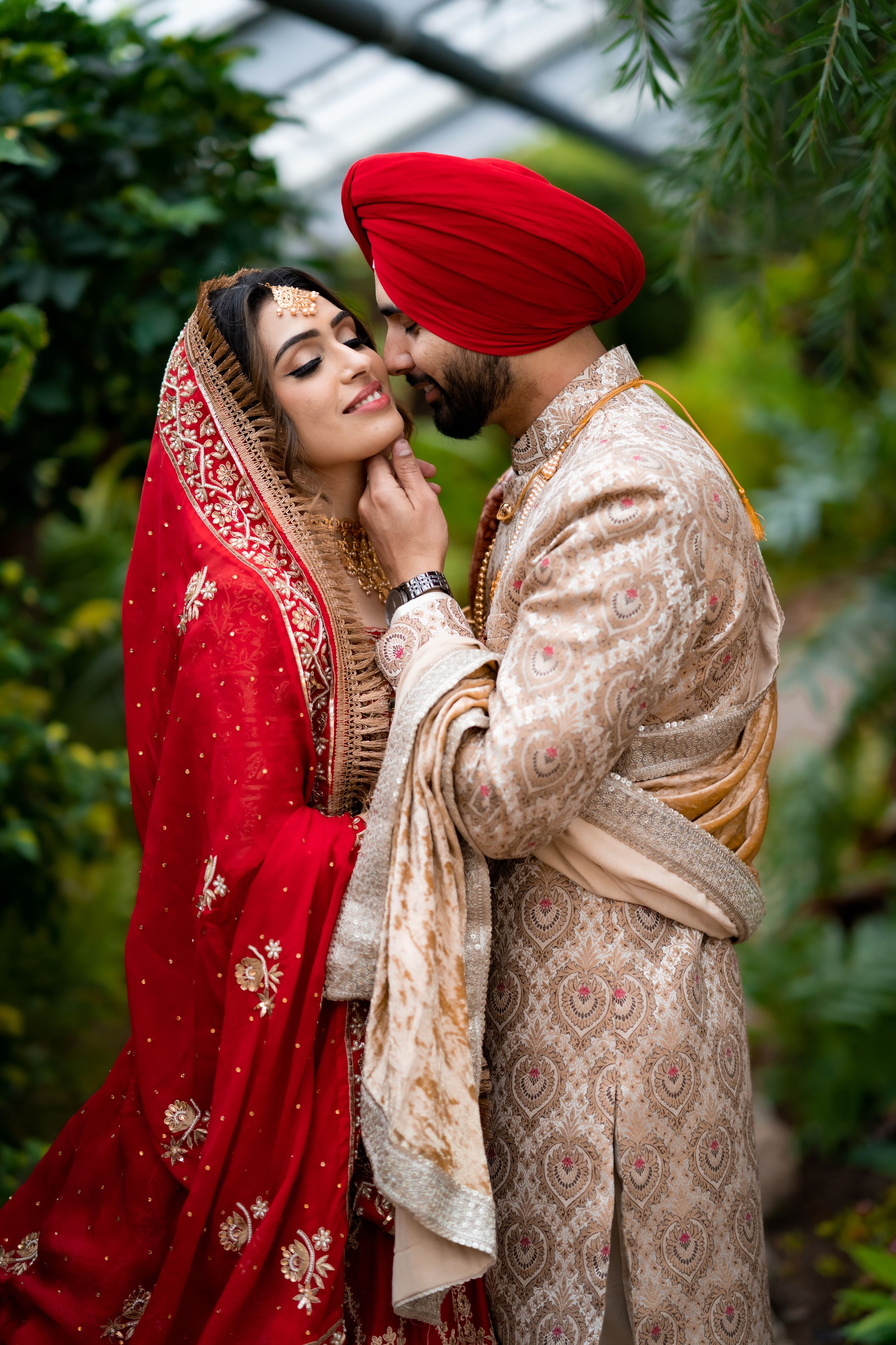 Traditional Sikh wedding couple in red and cream ornate attire share intimate moment in garden.