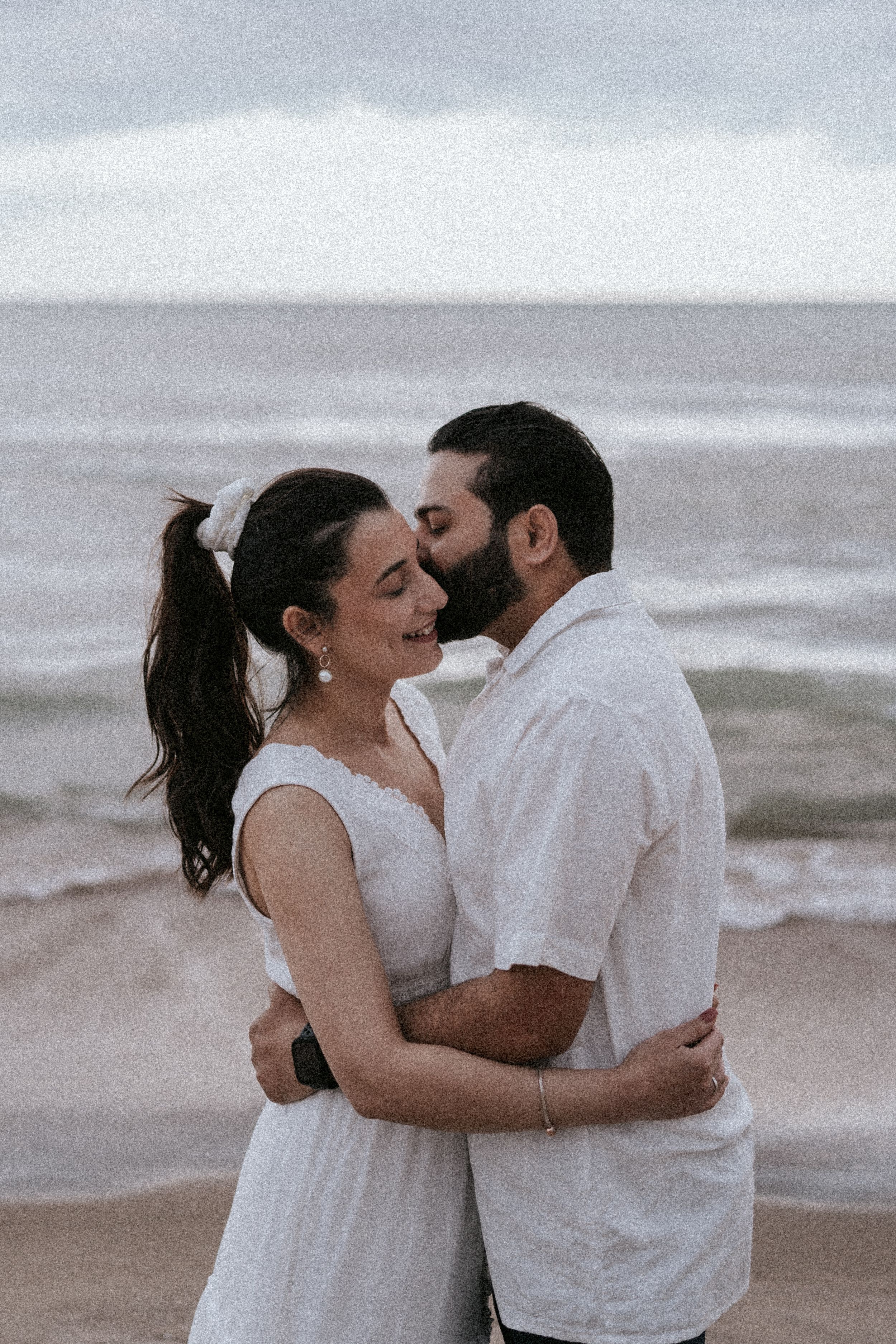Intimate beach portrait of couple embracing in casual white attire against ocean view.