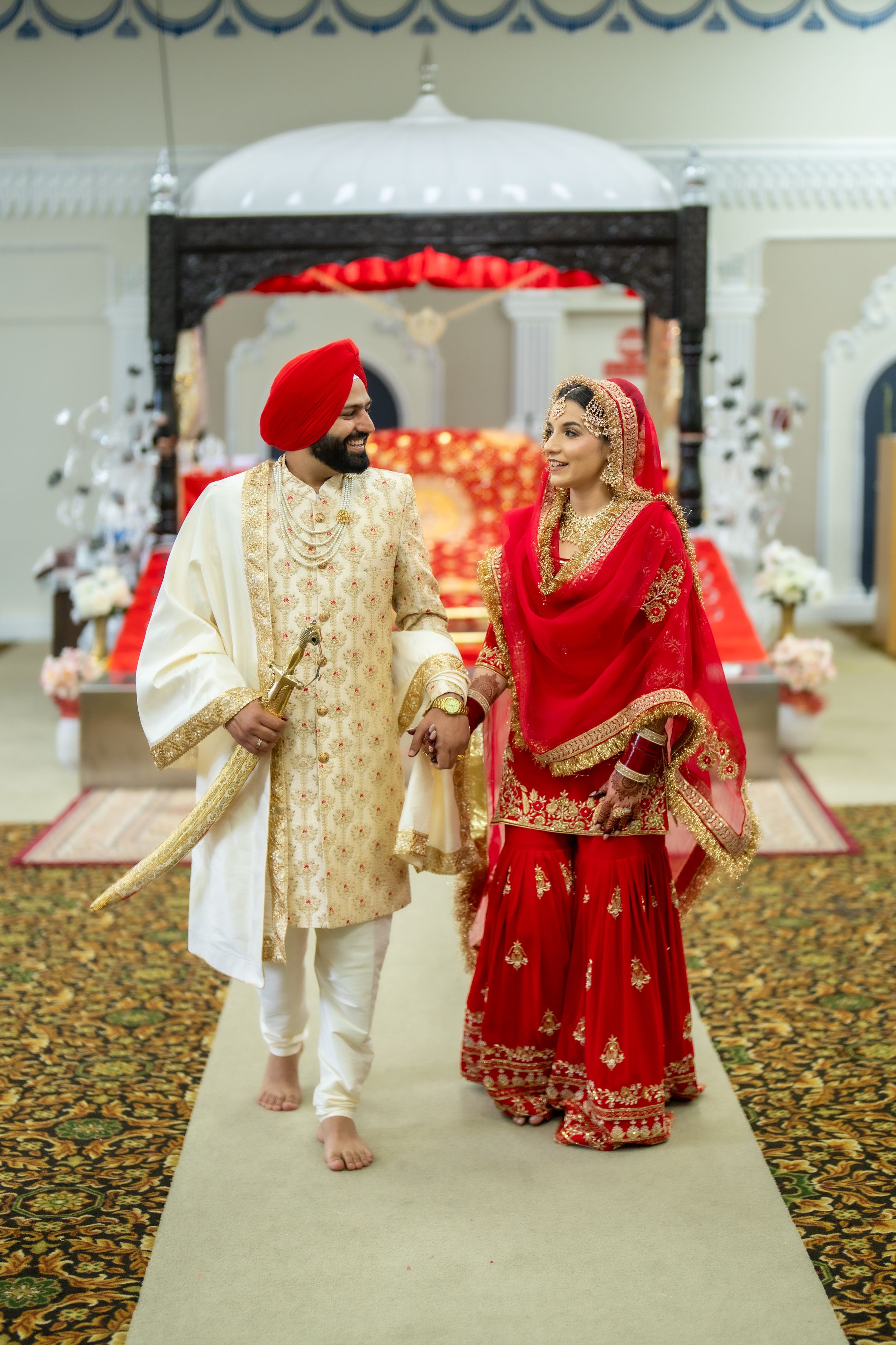 Traditional Sikh wedding couple in ornate red and cream attire walking down decorated aisle.