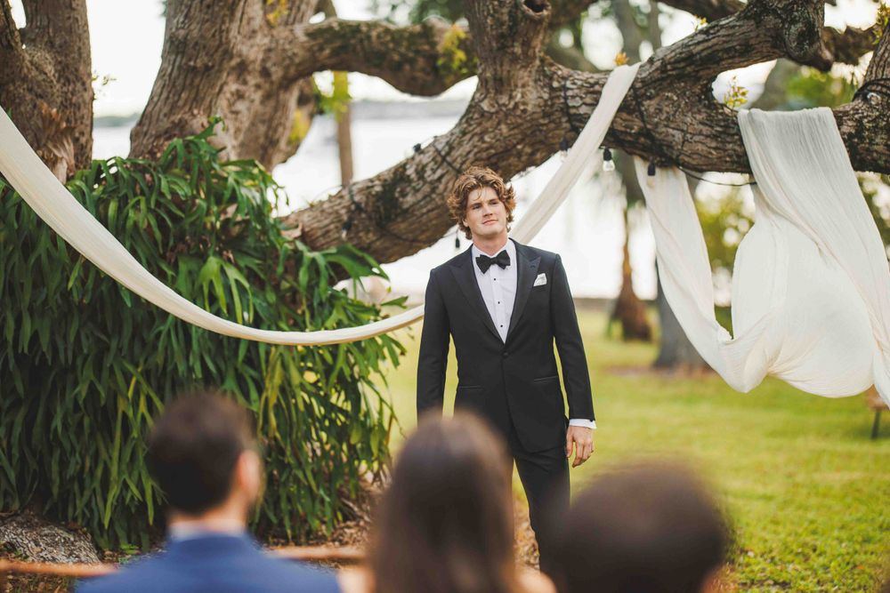 Groom in classic tuxedo standing under white fabric draped trees at outdoor ceremony.