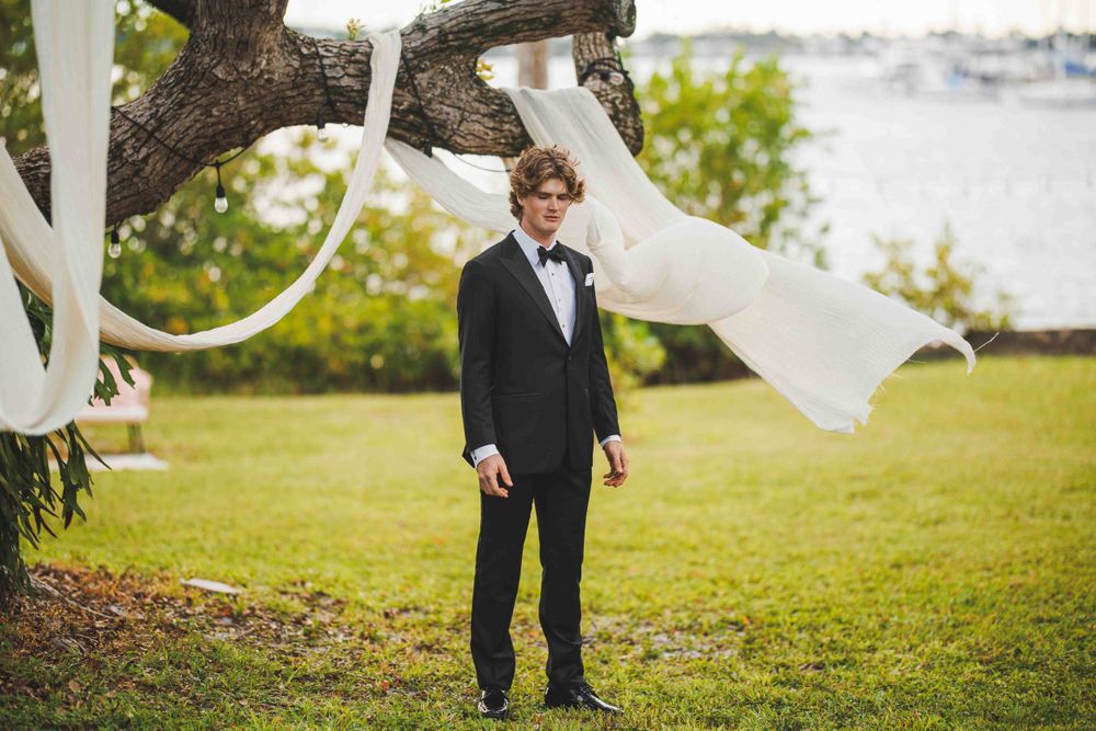 Groom in black tuxedo standing outdoors with flowing white fabric decorations in trees.