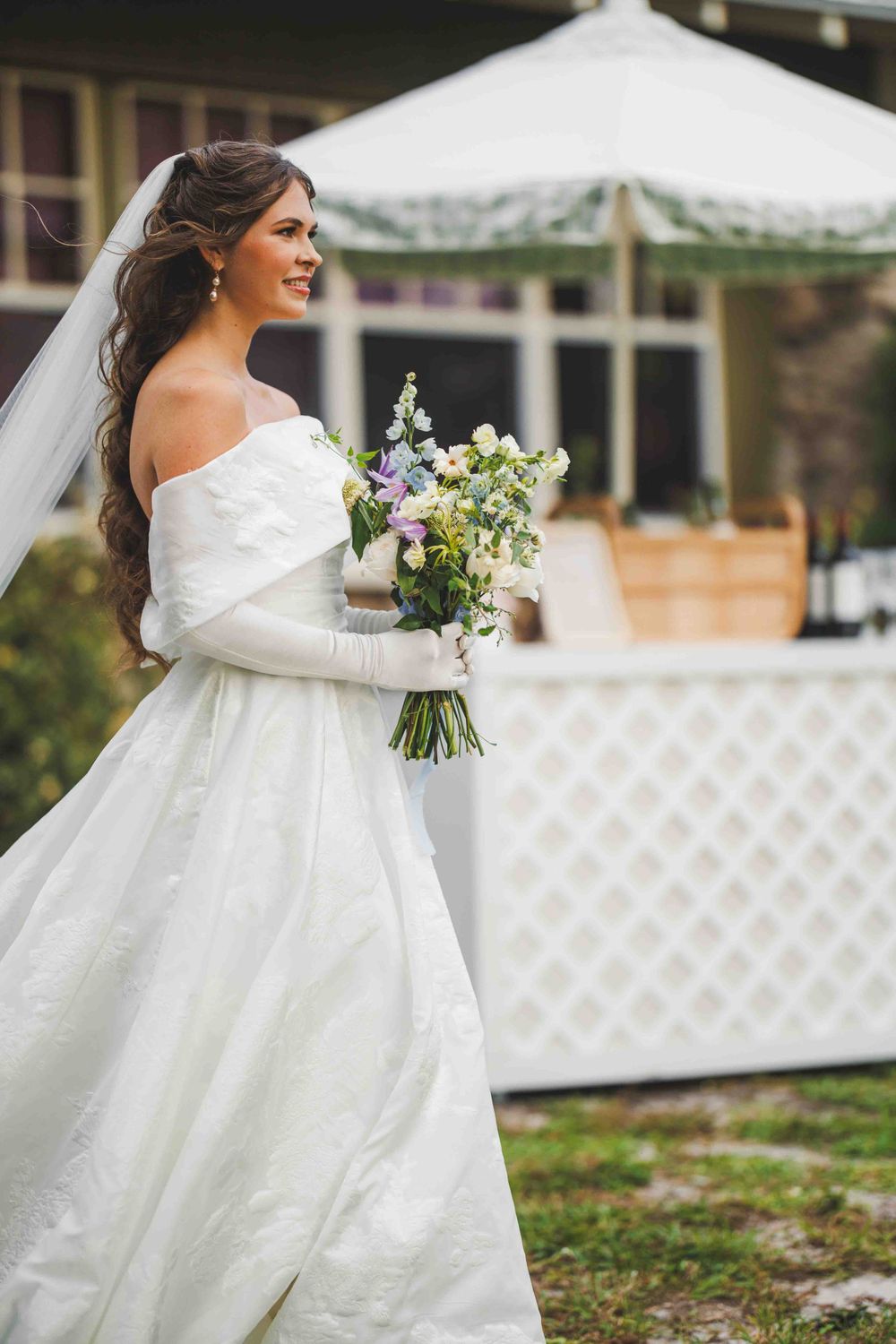 Bride in off-shoulder white gown holding a wildflower bouquet outdoors.