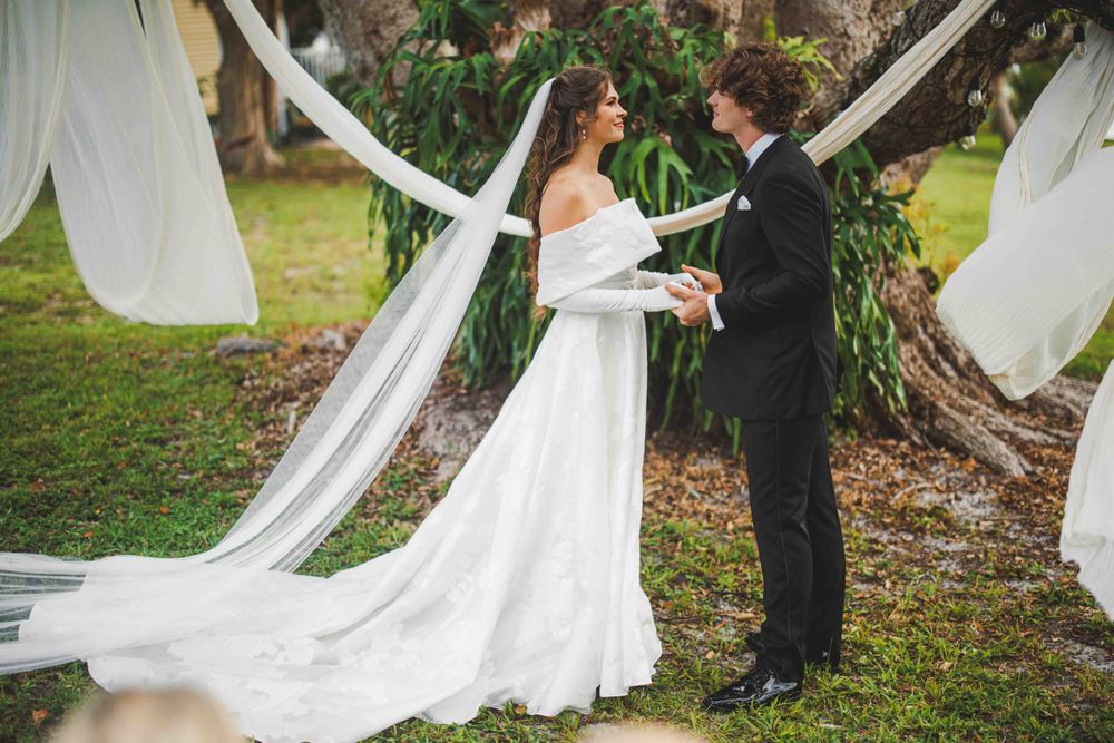 A wedding couple shares an intimate moment surrounded by flowing white fabric and greenery.