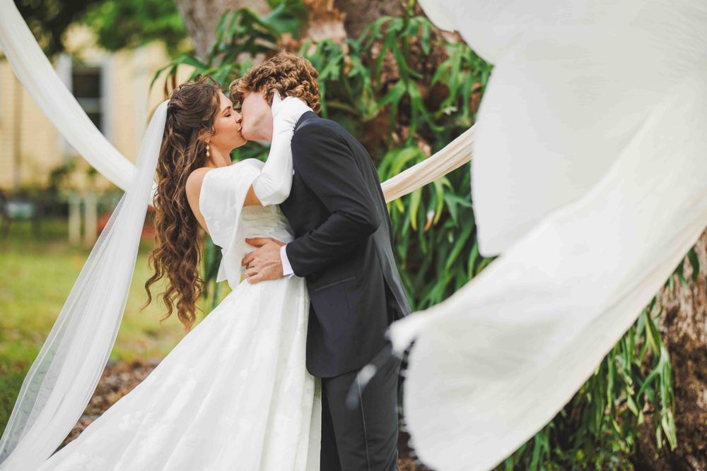 A romantic wedding kiss captured among billowing white fabric decorations outdoors.