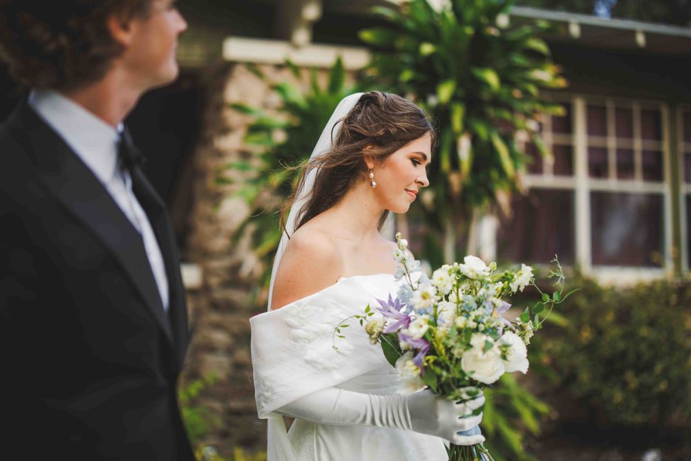 Wedding couple stands in a garden setting with elegant white floral bouquet.