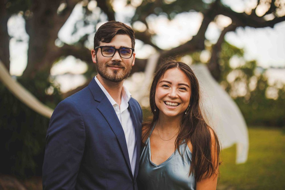 Couple smiles at camera with white fabric decor and trees in background.