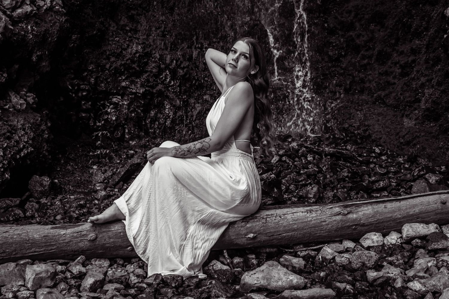 A black and white portrait of someone in a white dress posing on a log by stone steps.