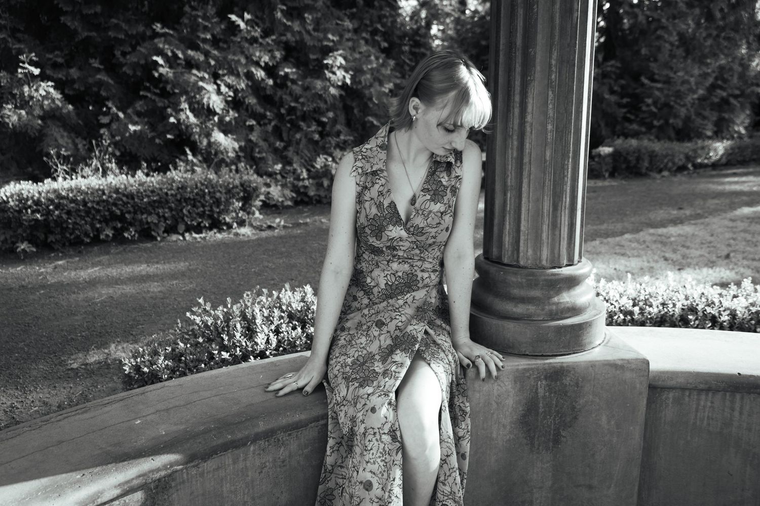 A vintage-style black and white portrait of someone in a floral dress seated by a classical column.
