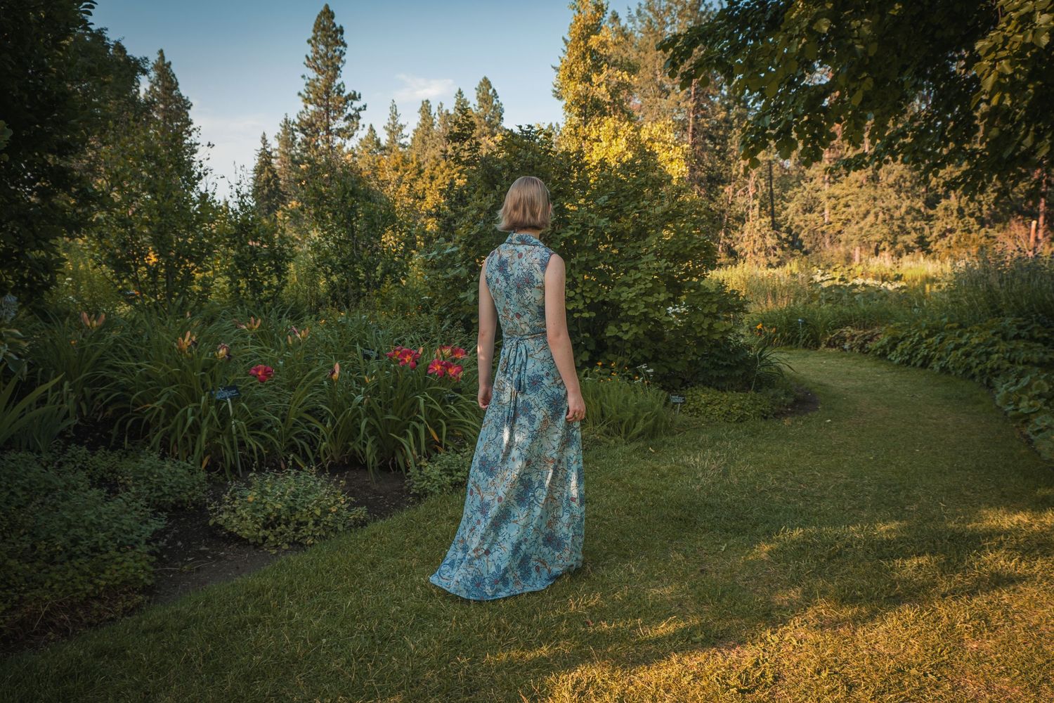 A floral blue maxi dress catches golden sunlight in a garden setting with evergreen trees and red flowers.