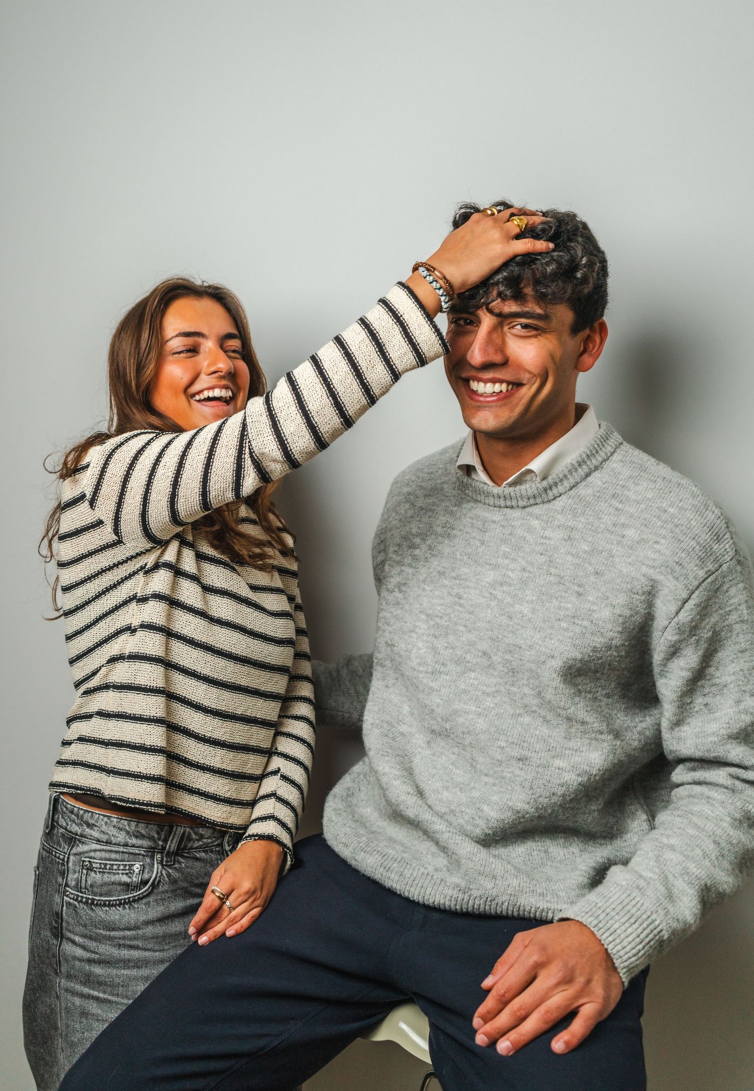 Two people in casual sweaters share a playful moment against a plain wall.