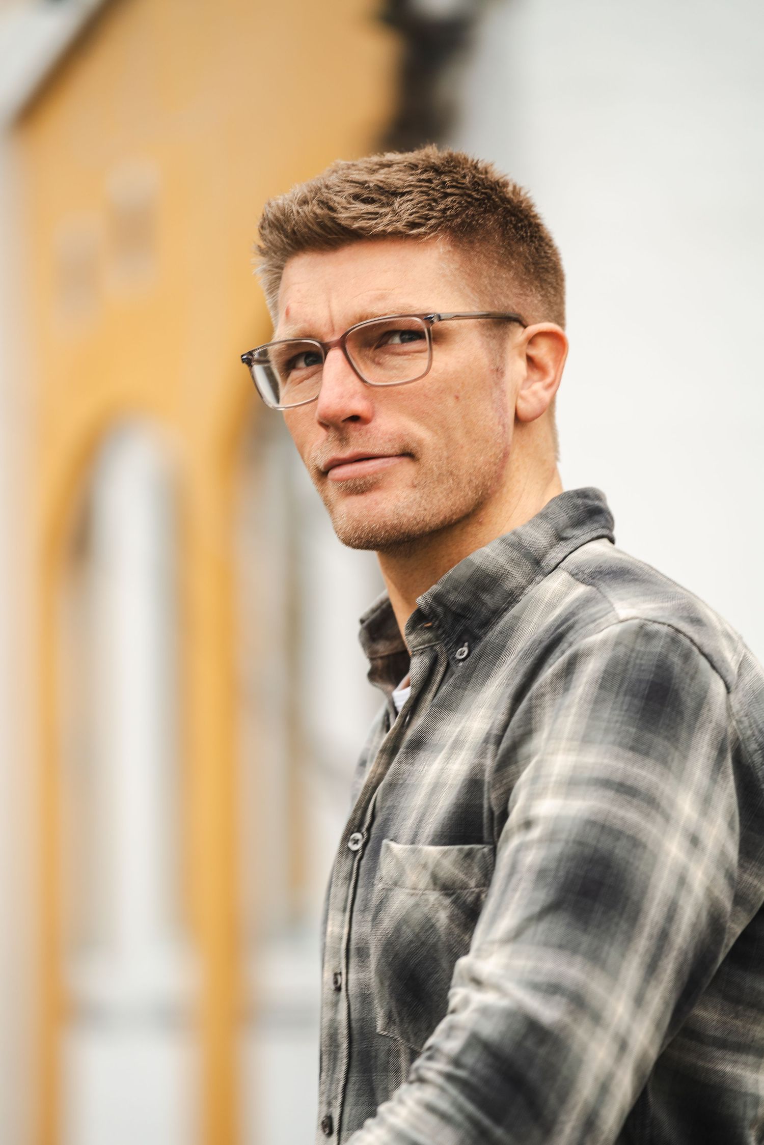 Casual outdoor portrait shows person wearing glasses and plaid flannel shirt against blurred yellow wall background.
