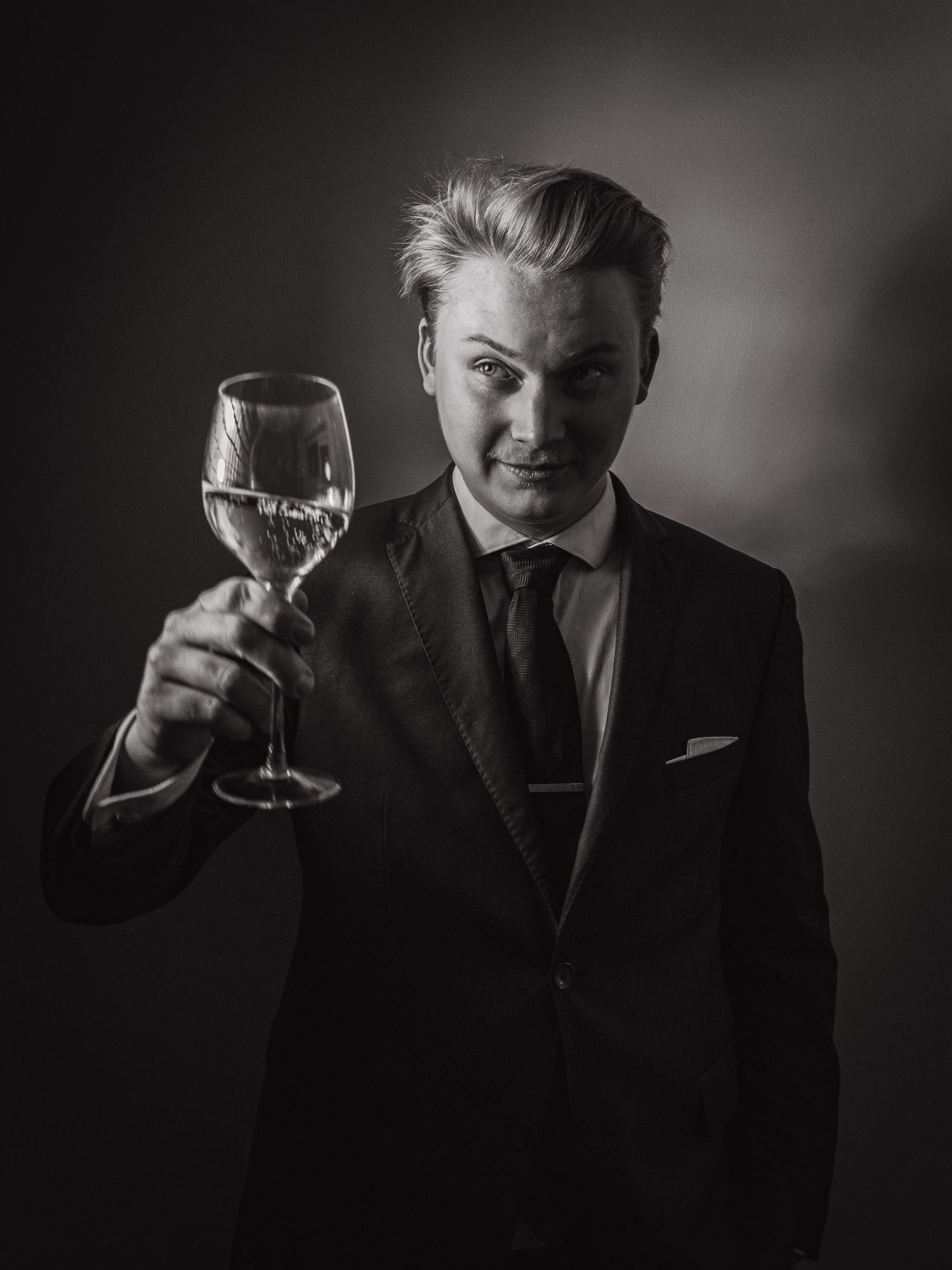Dramatic portrait of person in suit holding wine glass against dark background.