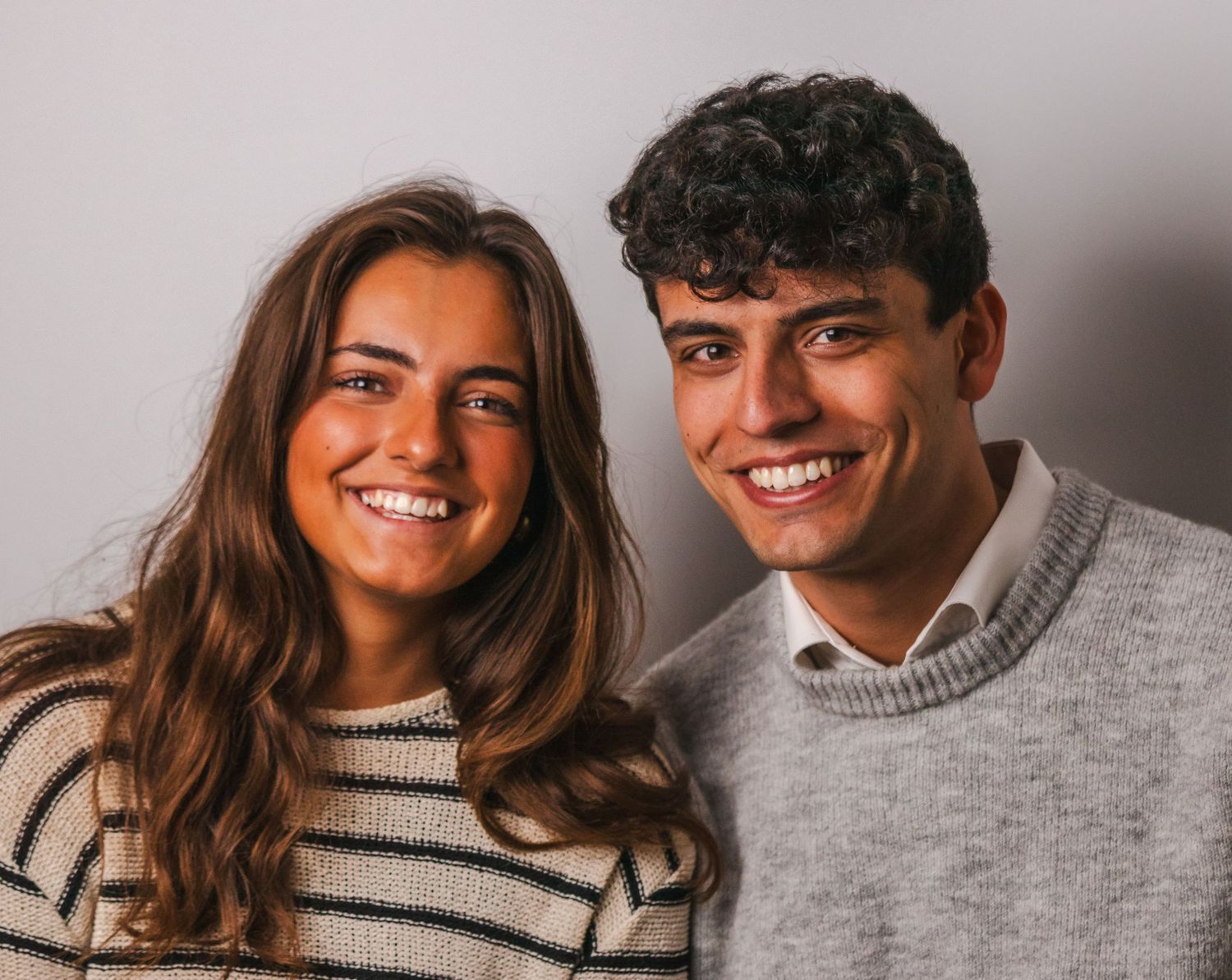 Two people in sweaters pose close together against a neutral background.