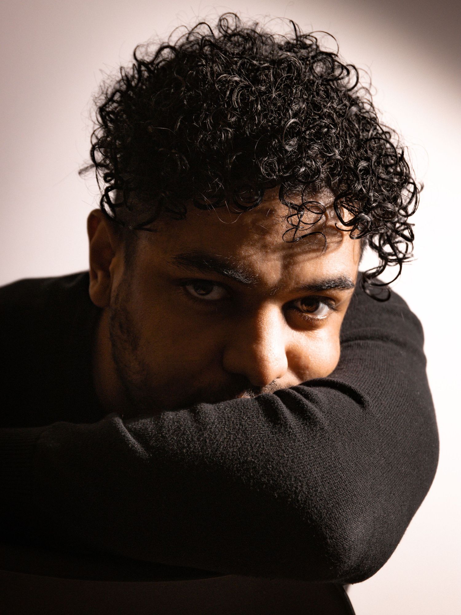 Artistic portrait of a person with dark curly hair resting their head on their arm against a dark background.