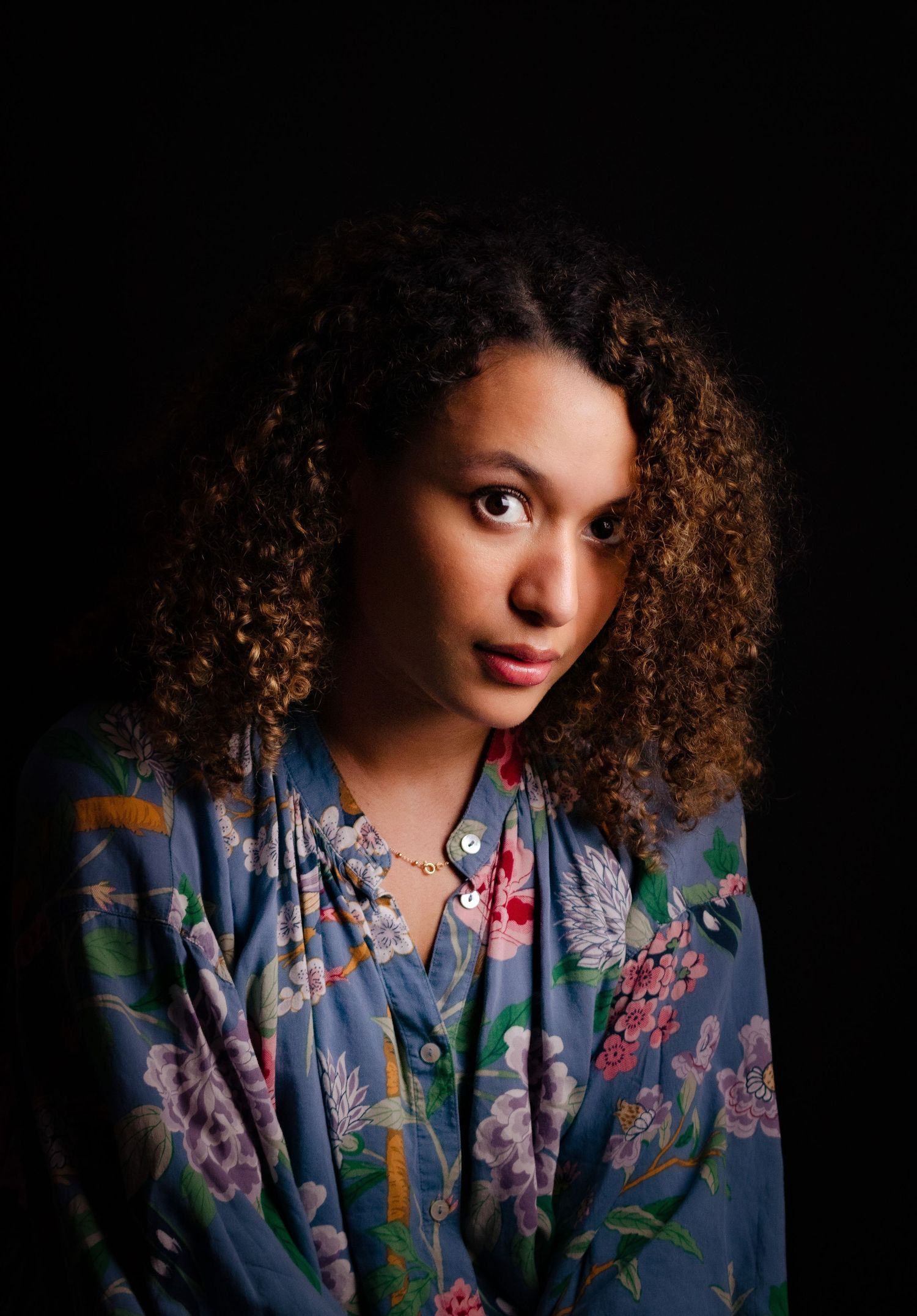Portrait of person in a floral dress against dark background with natural curly hair.