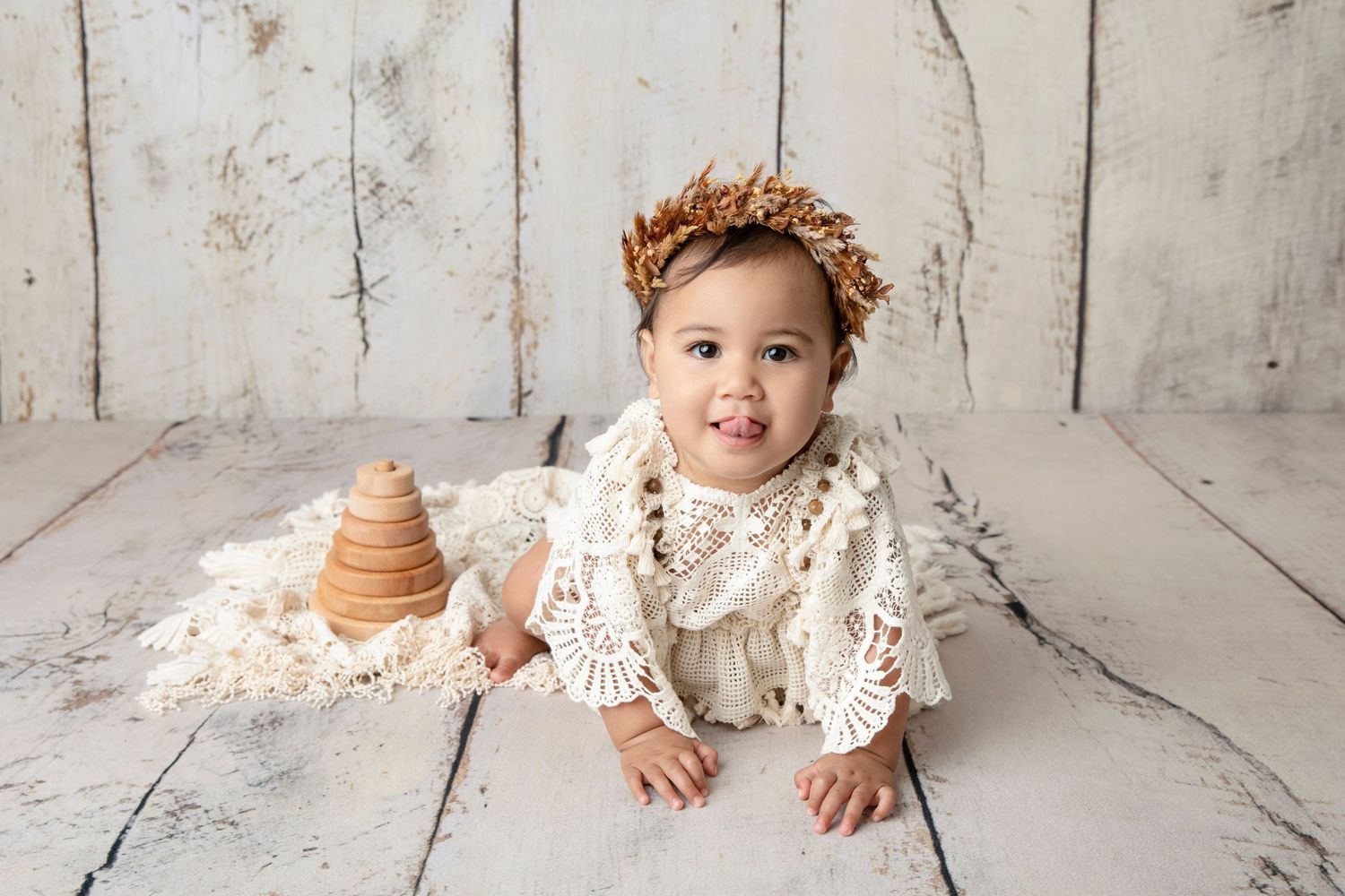 Baby in lace outfit sits beside wooden stacking toy against white background.