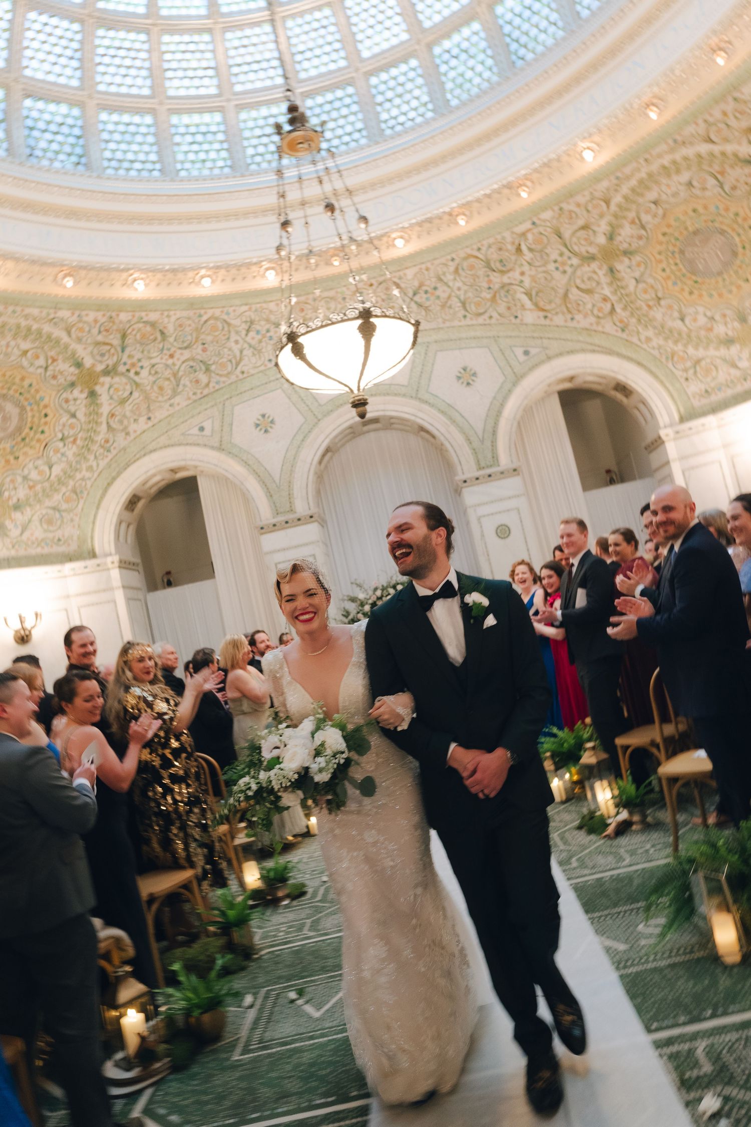 A bride in a white lace dress reads from a paper outdoors with joyful expression.