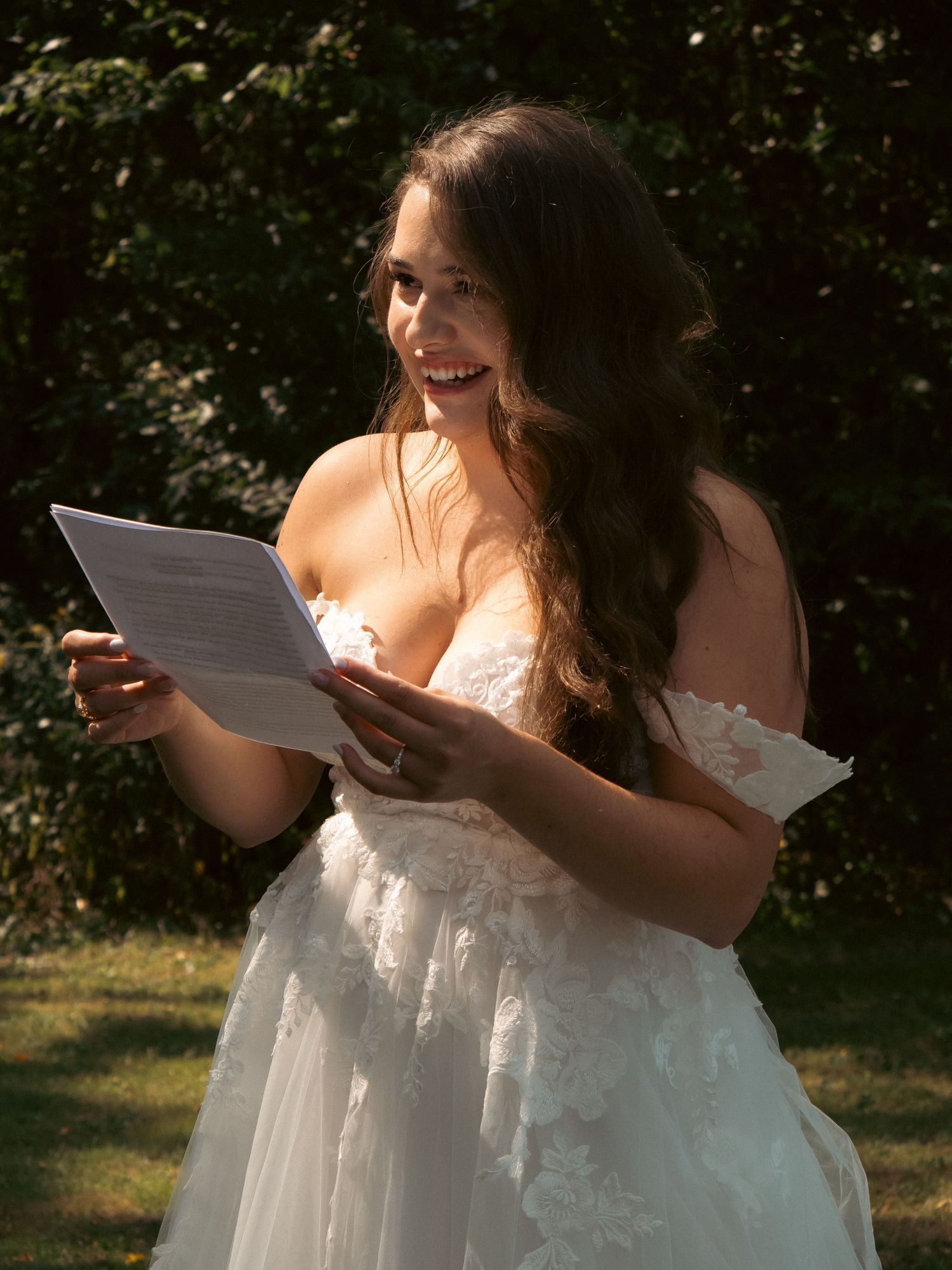 A bride in a white lace dress reads from a paper outdoors with joyful expression.