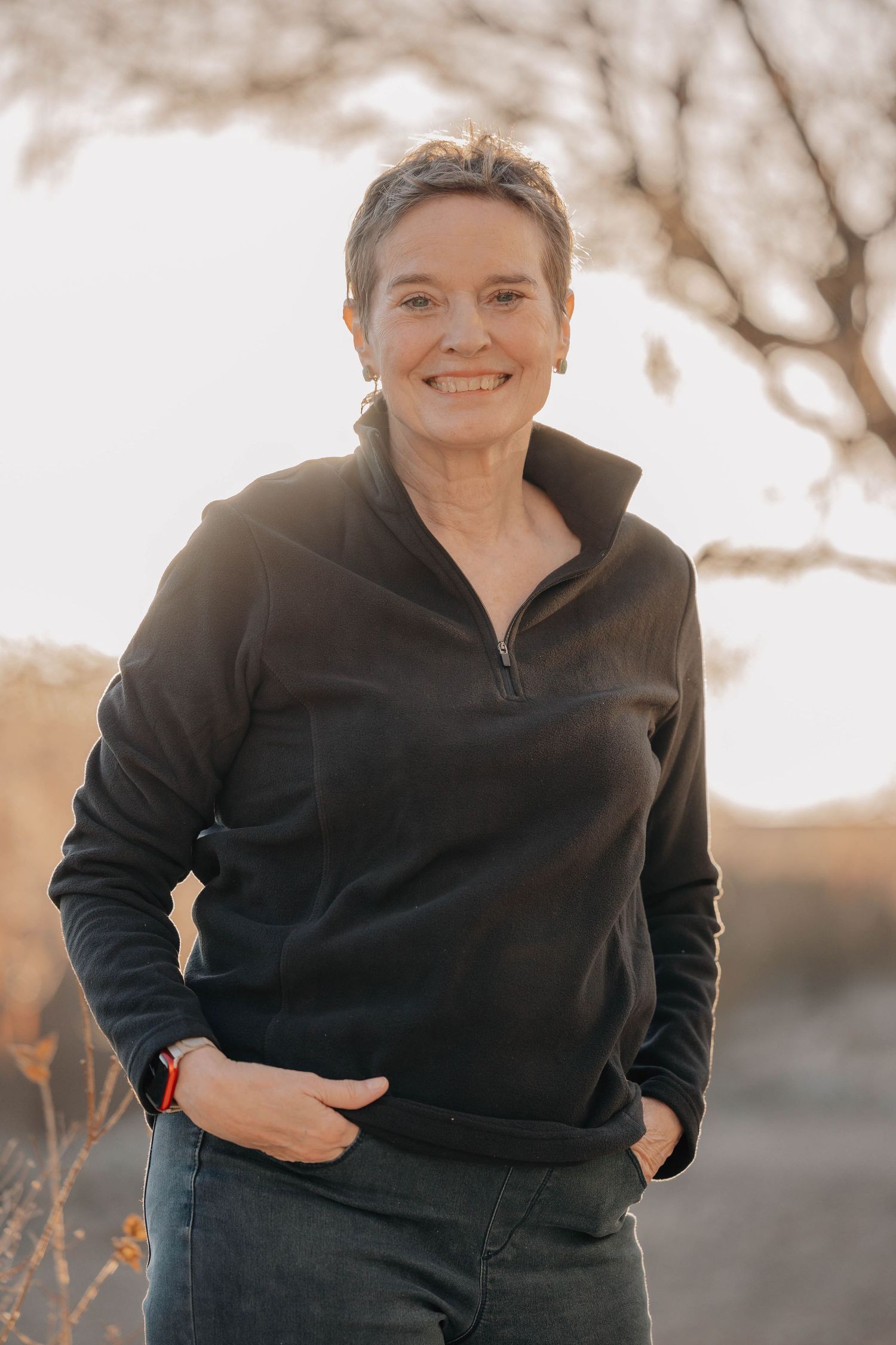 A person in a black quarter-zip pullover and jeans poses outdoors with hands in pockets during golden hour lighting.
