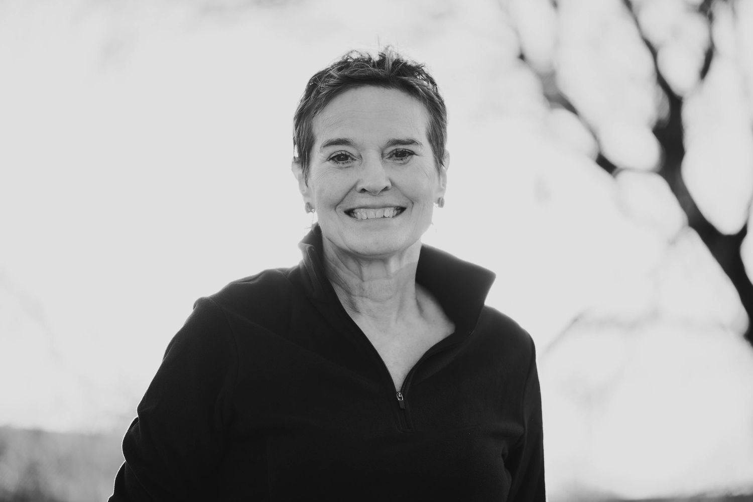 Black and white portrait of someone wearing a black collared top smiling warmly outdoors.