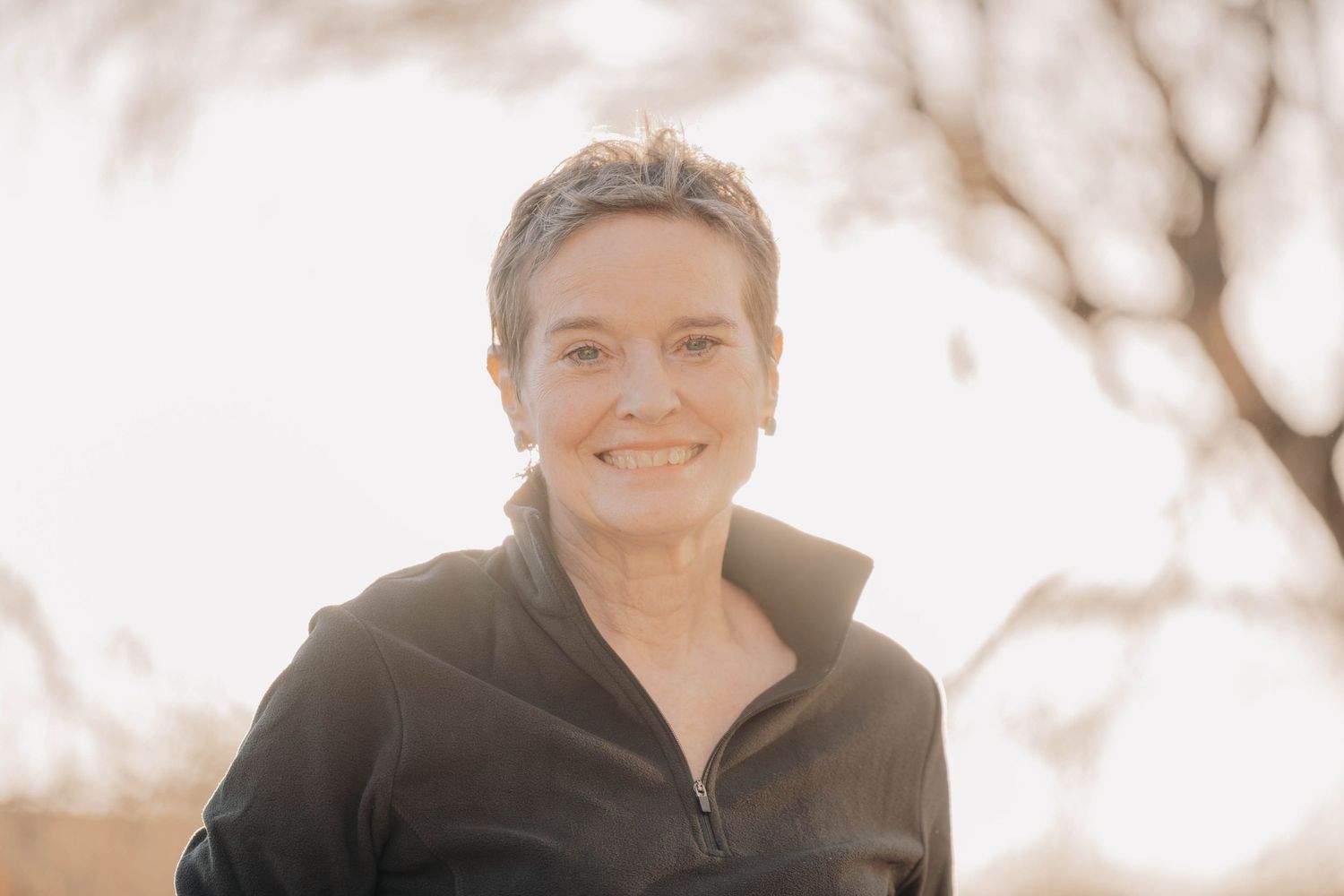 Person wearing a black fleece jacket smiles outdoors on a bright sunny day with soft bokeh background.