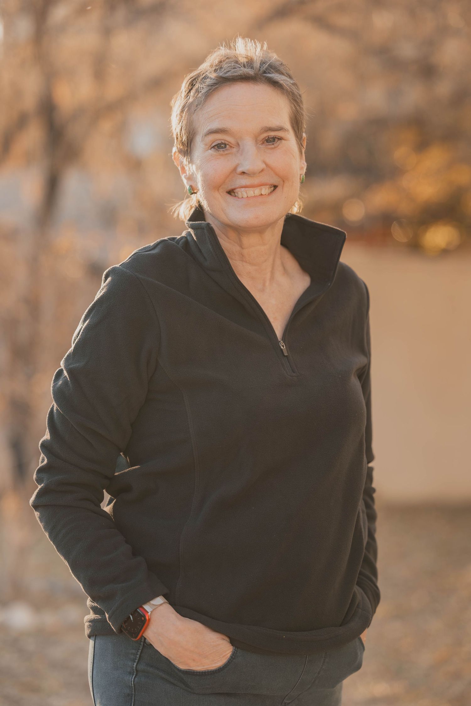 A person wearing a black quarter-zip sweater and jeans poses outdoors on an autumn day with warm lighting.