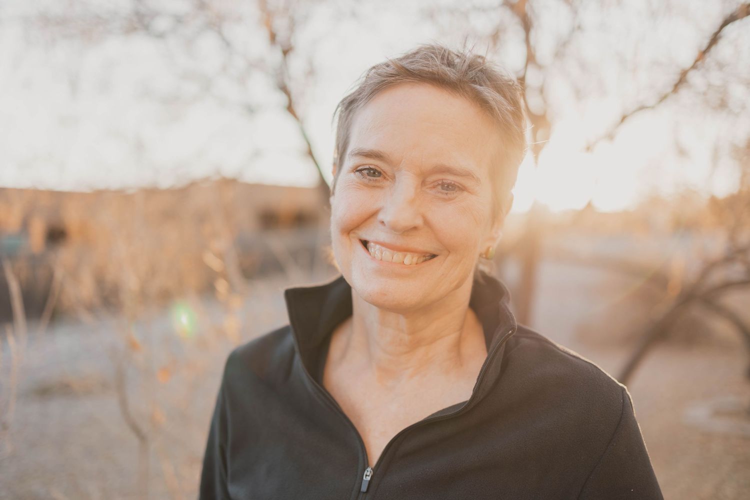 Person wearing black fleece jacket smiles warmly while standing outdoors against blurred autumn background.