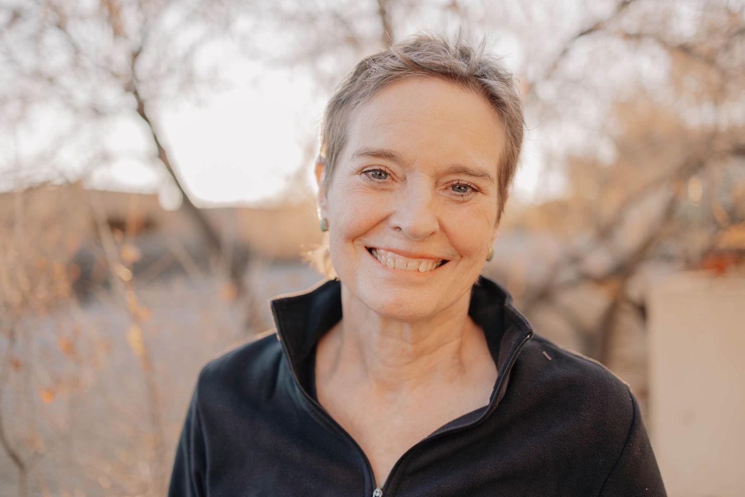 Person wearing black fleece jacket smiles outdoors with blurred autumn trees in background.