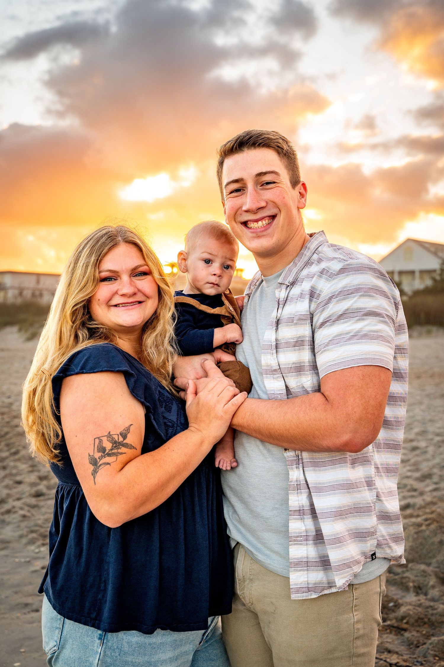 Family portrait on beach at sunset with vibrant orange sky and coastal homes in background.