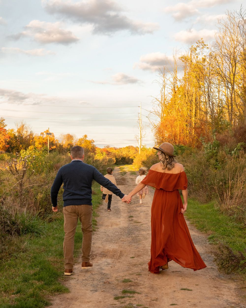 A couple walks hand in hand down a rustic path during golden hour with fall trees glowing in the background.
