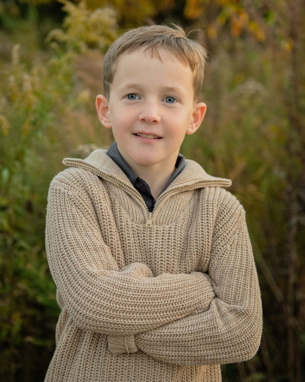 A young boy in a beige knit sweater poses outdoors against an autumn background.