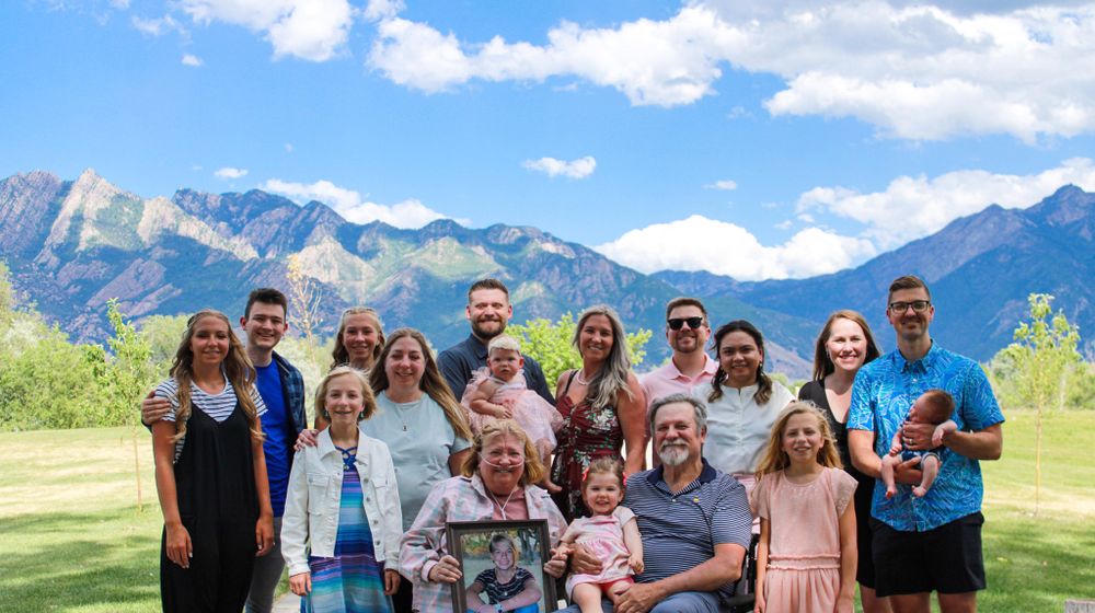 Large family gathering poses outdoors with mountains and blue sky in background.