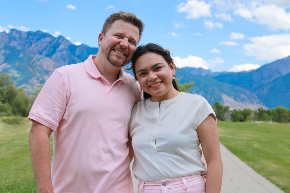 Couple in casual attire posing with mountain range backdrop on sunny day.