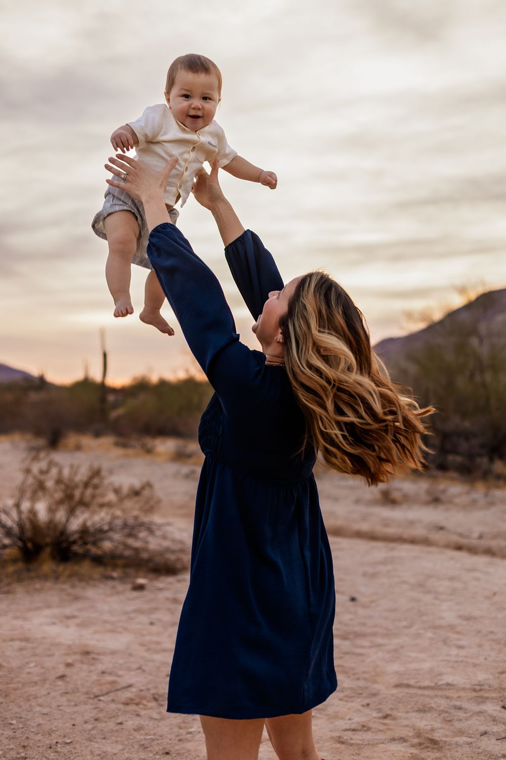 Woman in navy dress lifting baby high in air against desert sunset backdrop in Phoenix Arizona for her family photoshoot.