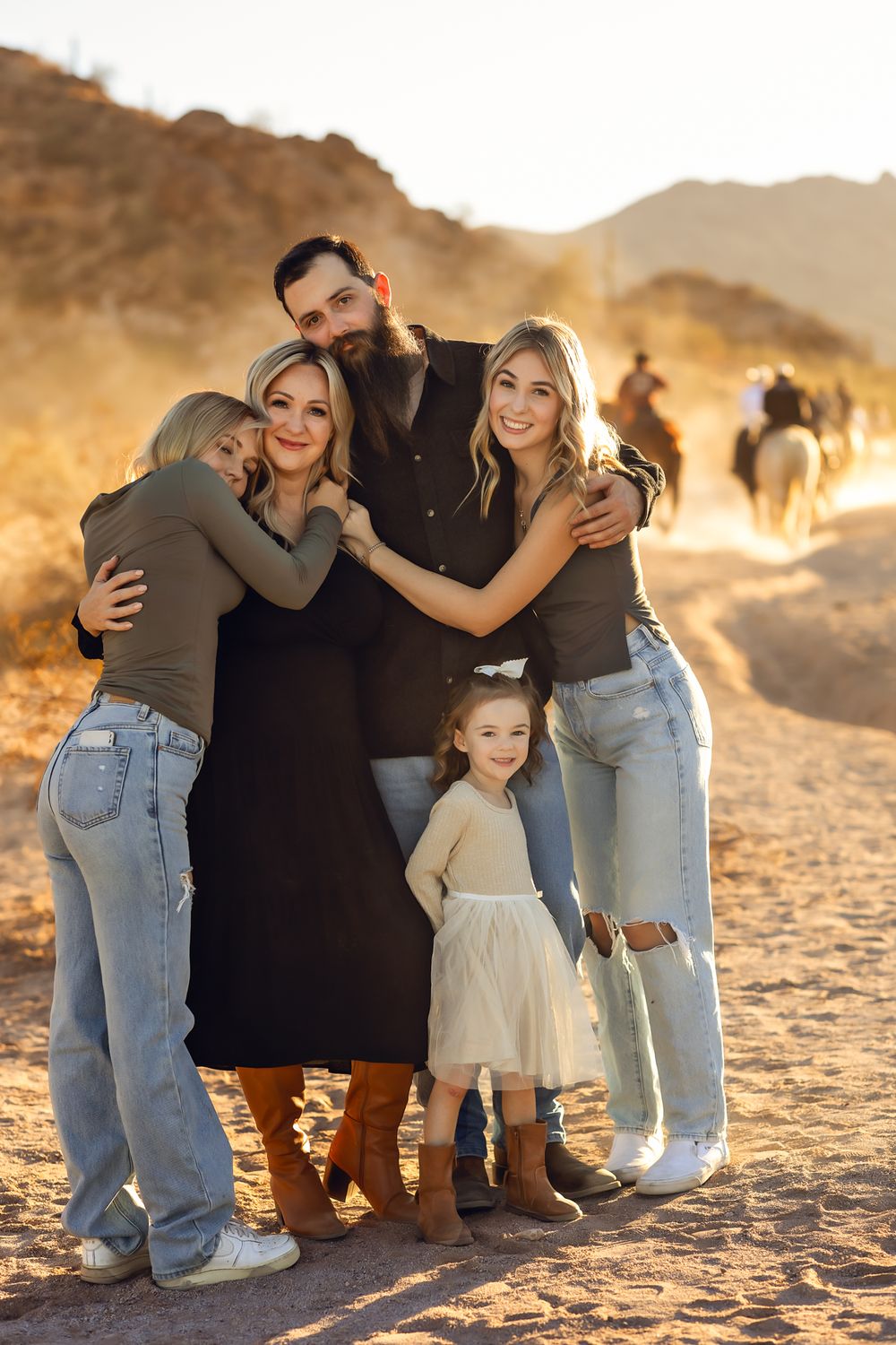 A family embraces during a desert photoshoot at golden hour with mountain scenery and horseback riders in the background for their photos