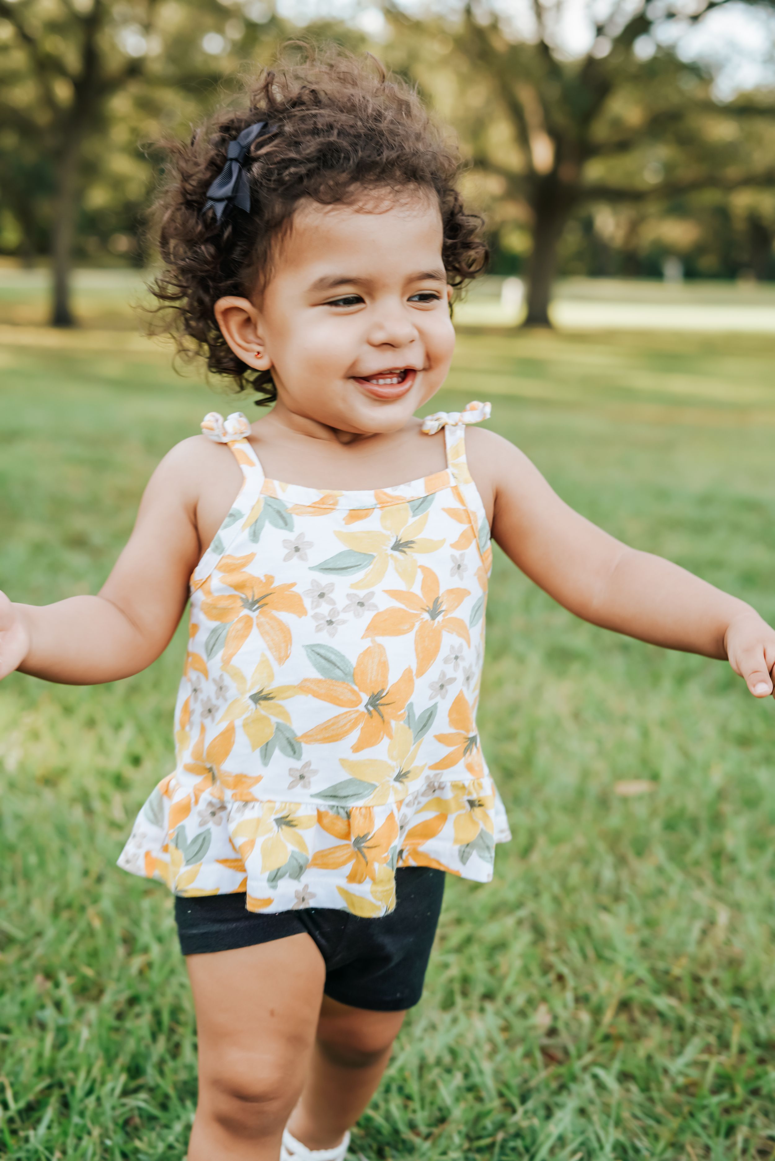 Toddler in floral print top and shorts plays joyfully on green grass.