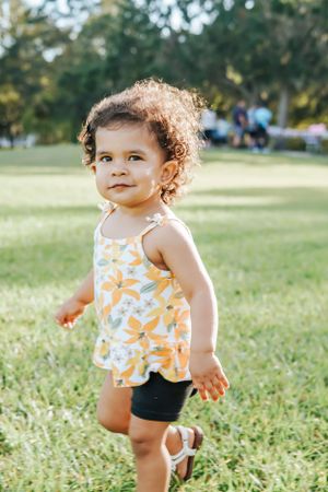 Toddler wearing orange floral top exploring sunny grassy field.
