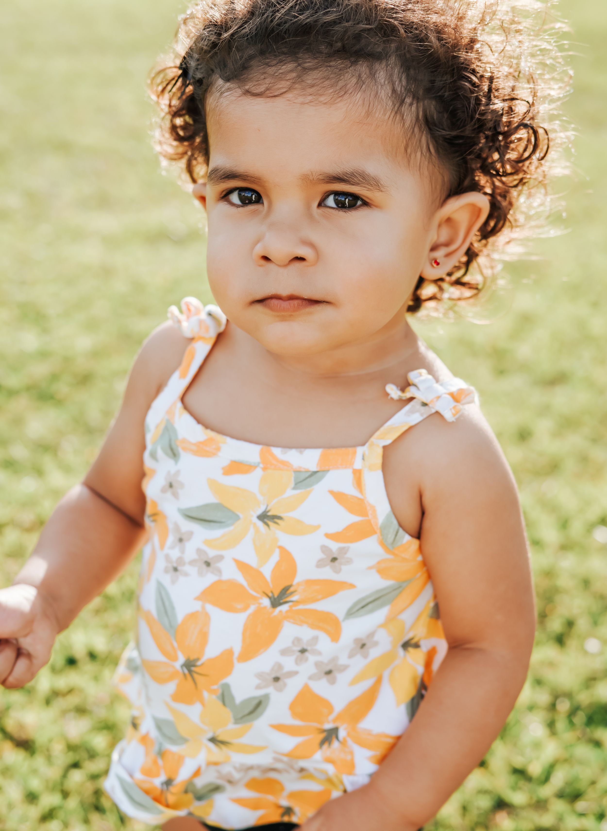 Baby in orange floral tank top sitting in sunlit grassy area.