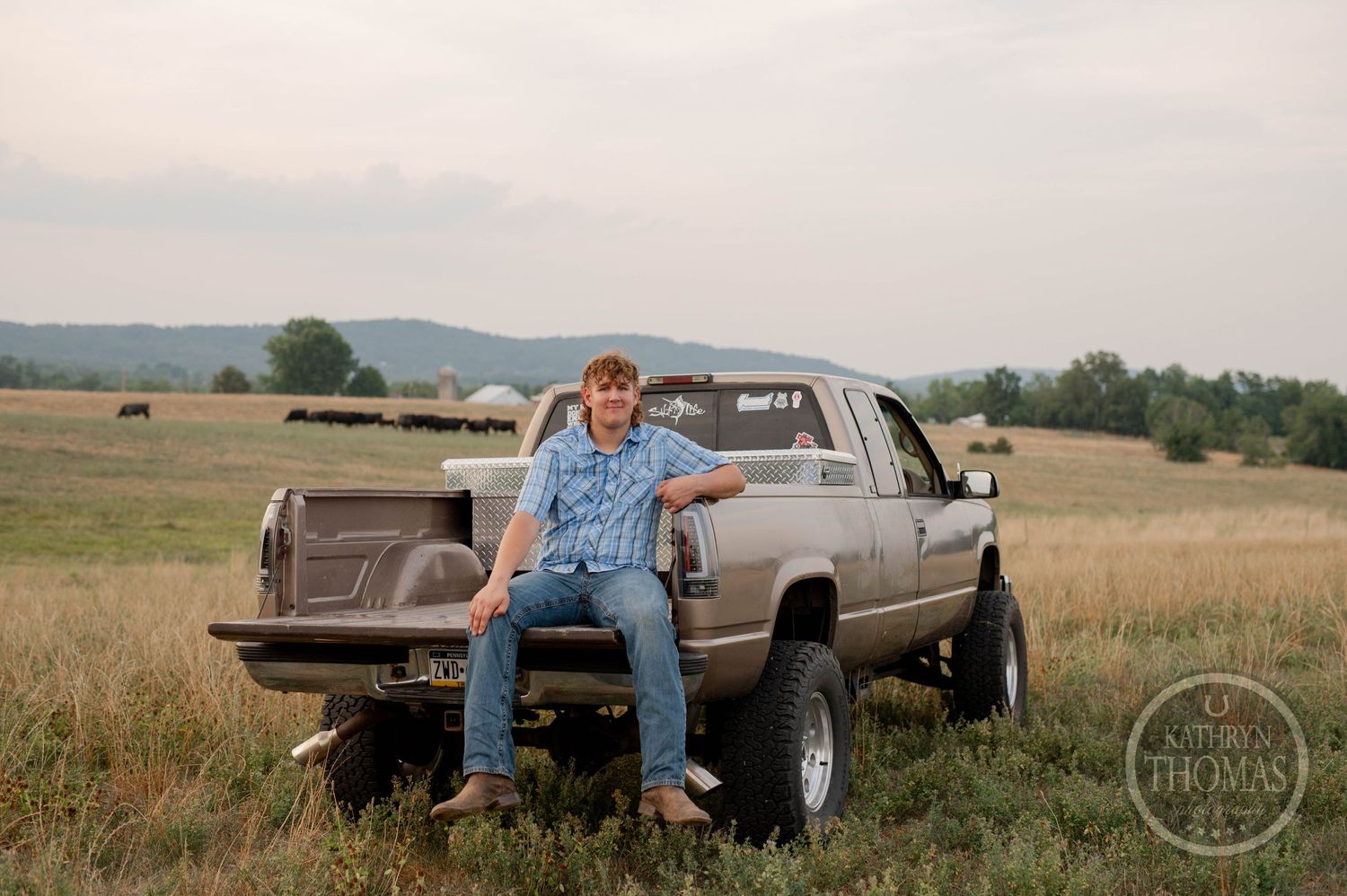 A person in blue plaid and denim poses on a lifted silver pickup truck in a rural field.