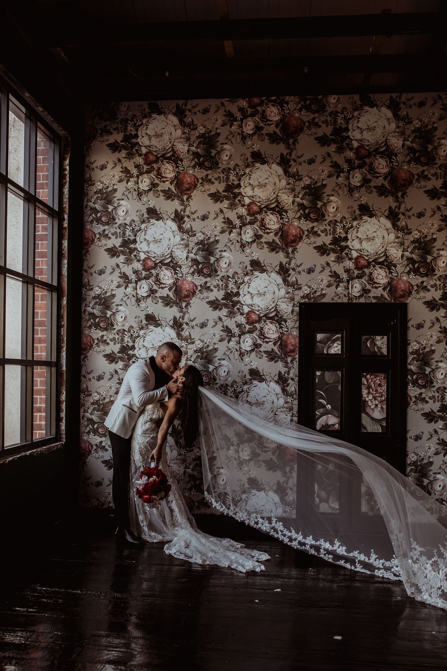 In a vintage room with floral wallpaper, a long flowing veil cascades from a wedding couple's romantic pose near tall windows.