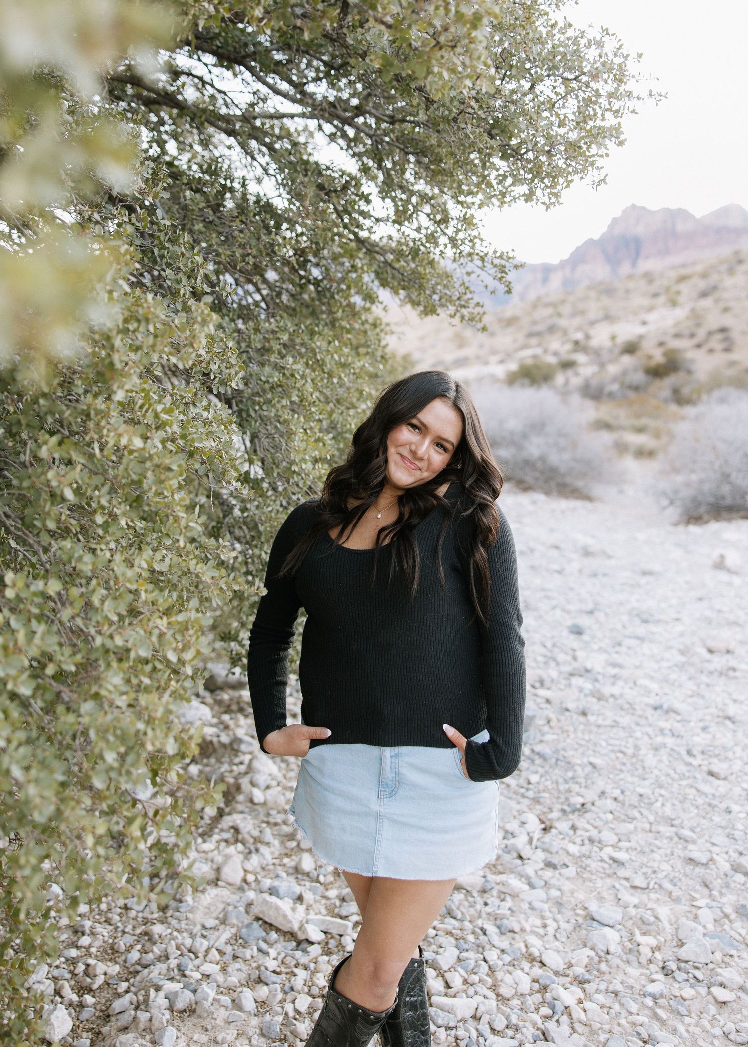 Person in black sweater and denim skirt poses on desert trail with mountains and desert foliage in background.