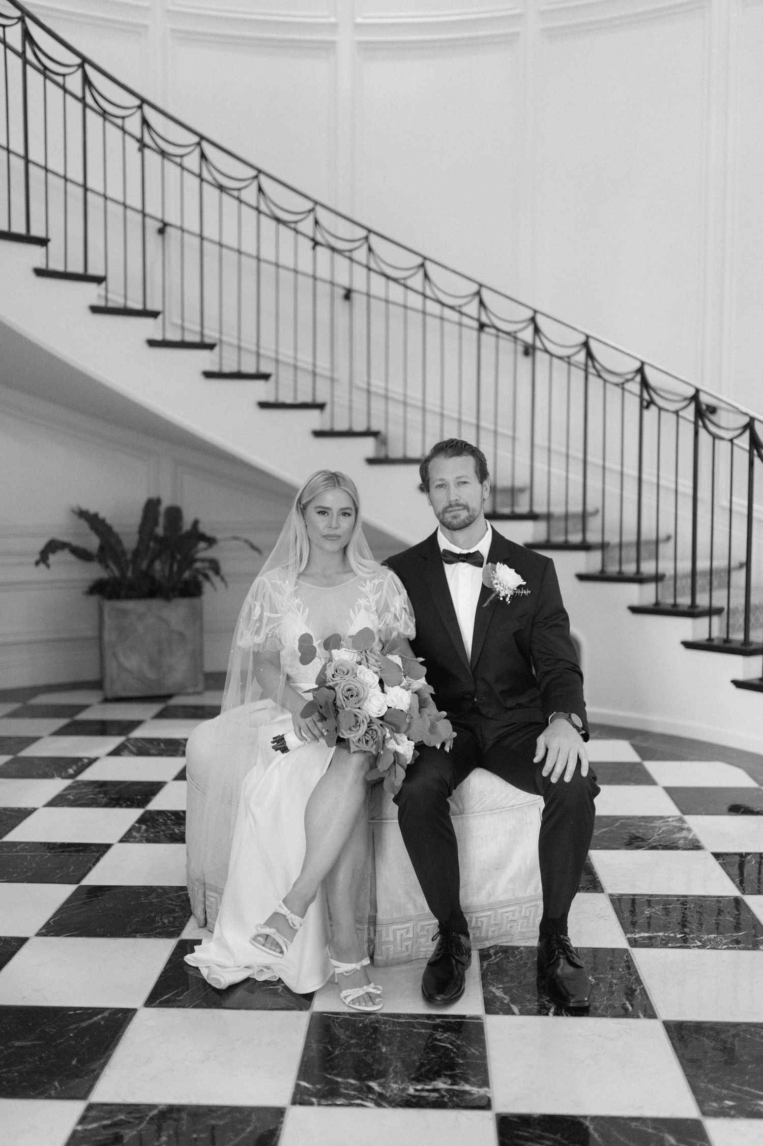 A wedding couple sits on a black and white checkered floor in front of an elegant curved staircase.