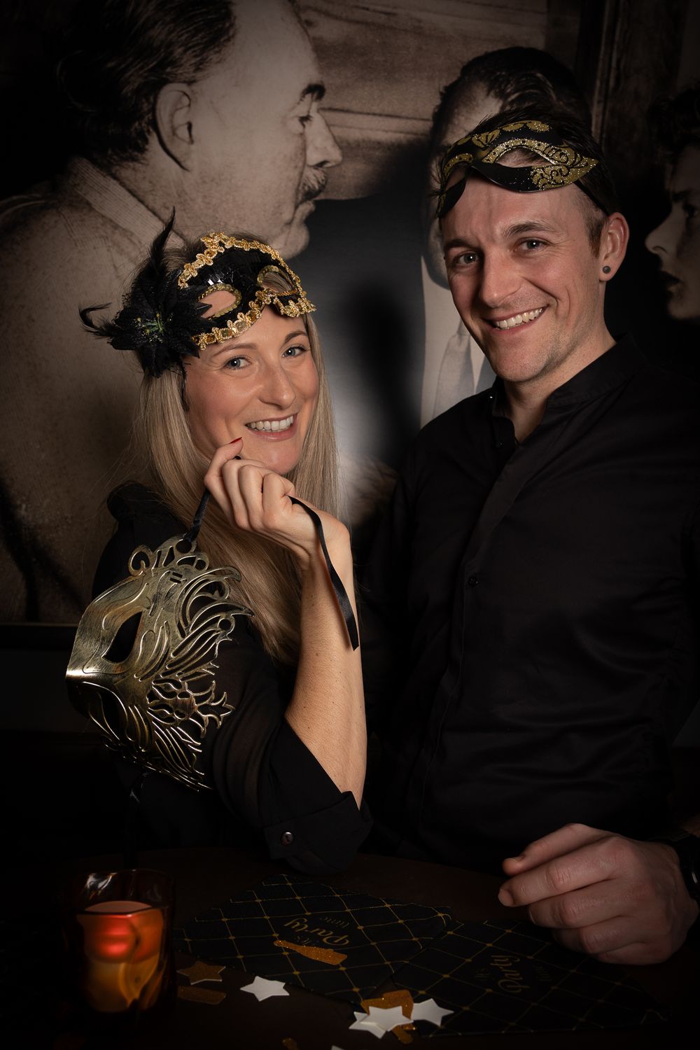 Two people wearing elegant black outfits and ornate gold headpieces pose together in a dimly lit vintage-style portrait setting.