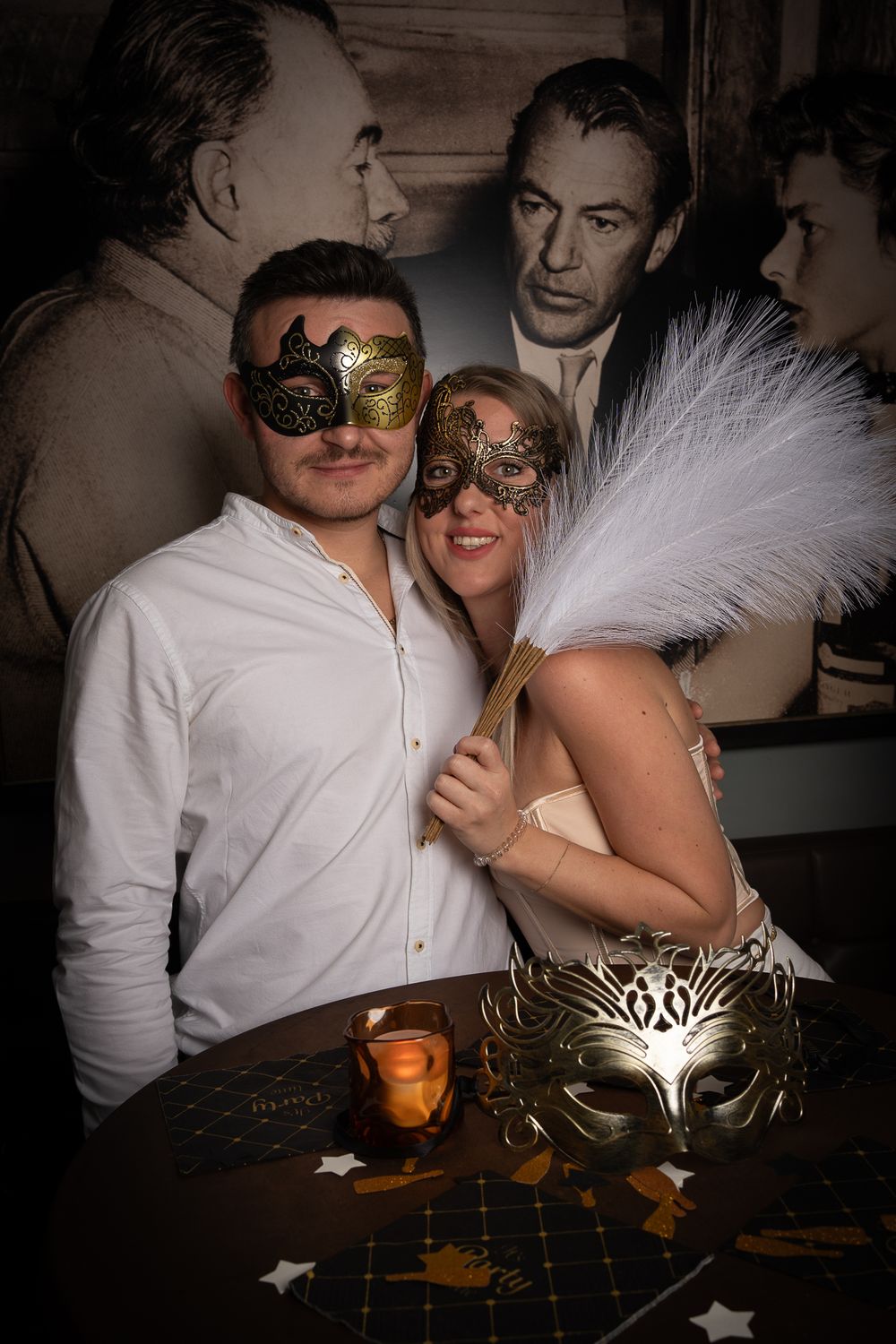 A couple wearing ornate masquerade masks pose together at an elegant ball, with decorative feathers and masks on display.