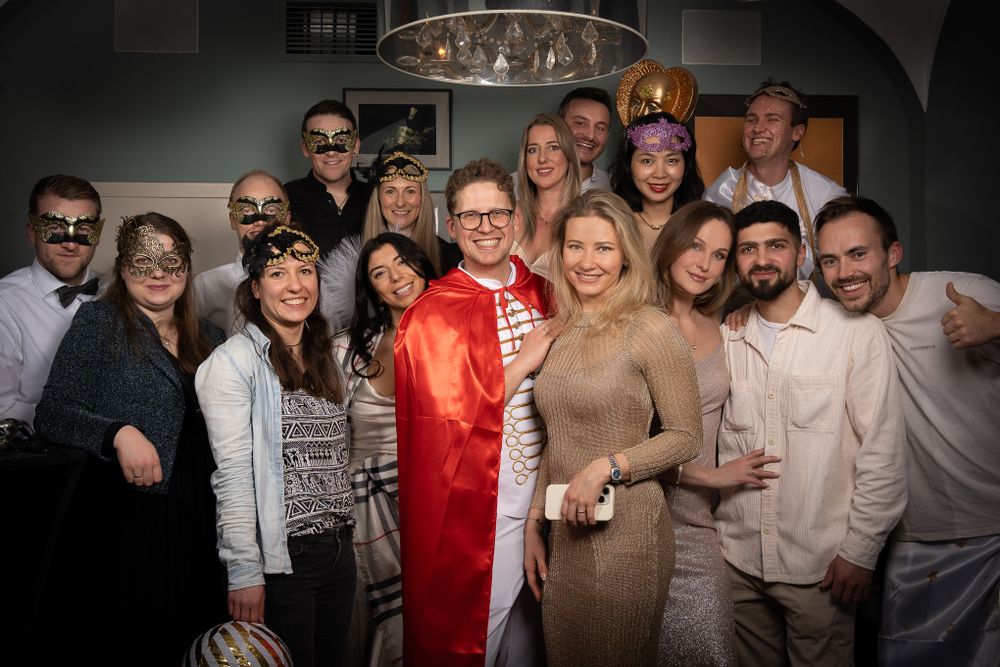 A group of people dressed in casual and formal attire pose together at an indoor social gathering under dim lighting.
