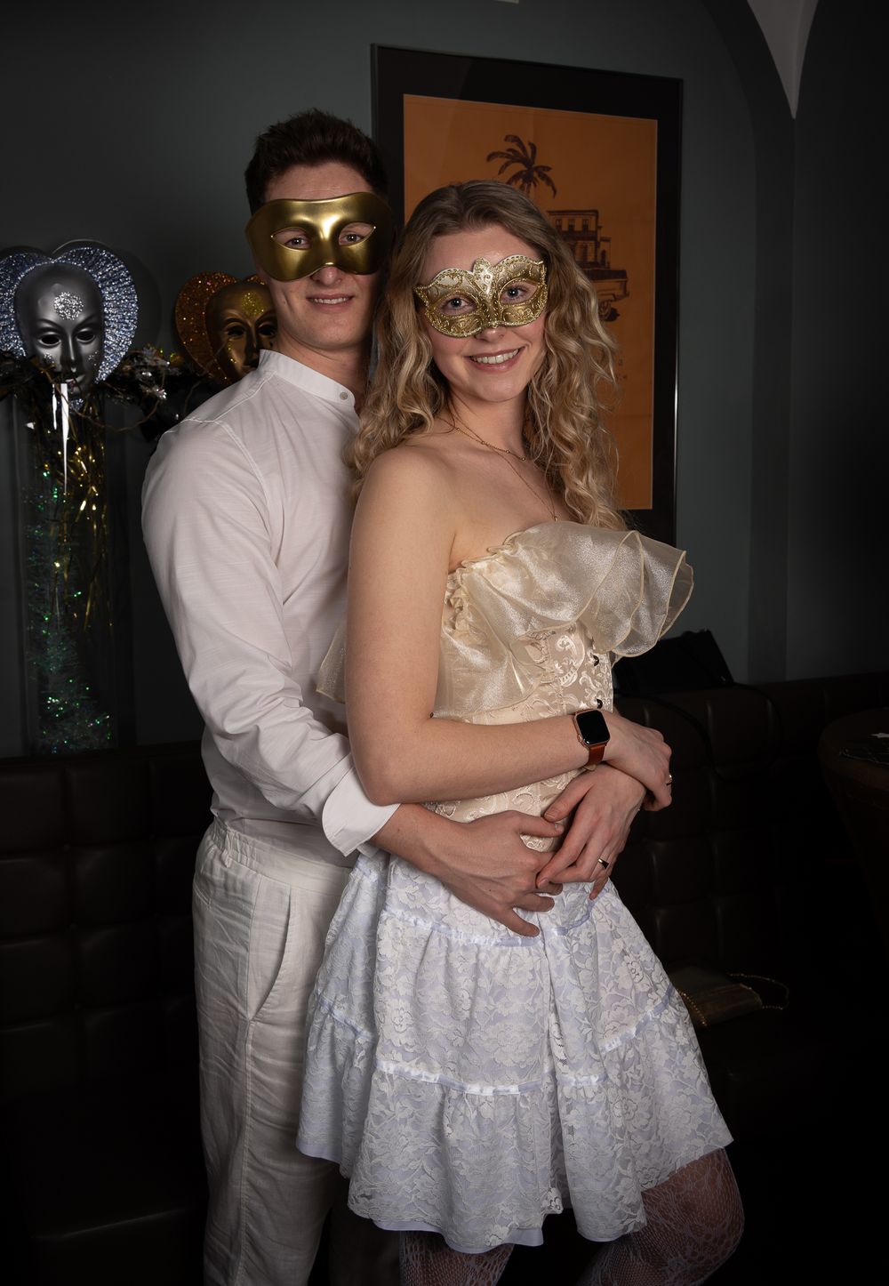 A couple in formal attire wearing ornate masquerade masks pose together at a party.