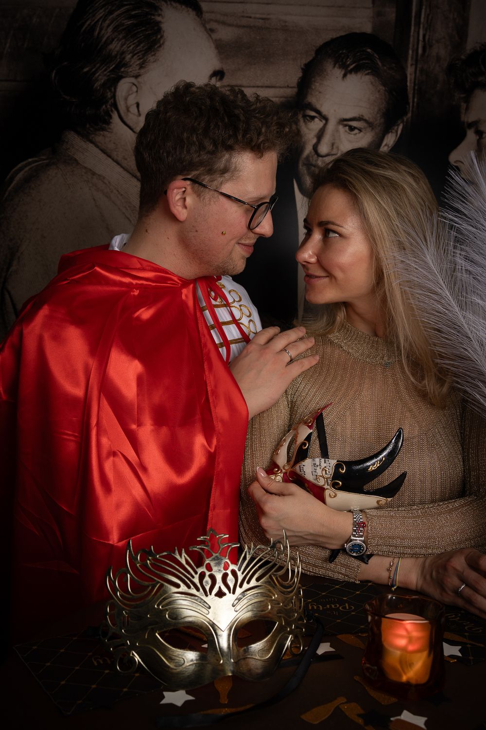 Couple in festive attire share an intimate moment at a masquerade party with decorative mask and candle on table.